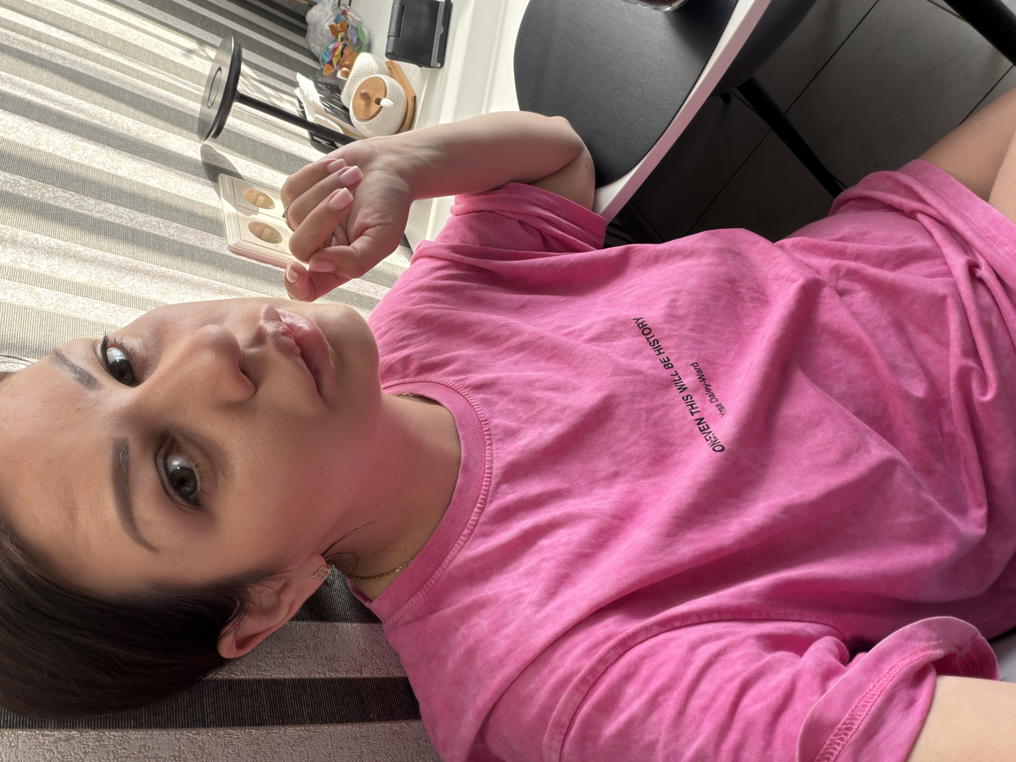 A woman with dark hair is sitting at a desk in a room, possibly her home office. She is wearing a pink t-shirt with text on it. The room has striped wallpaper and a window with blinds. On the desk are some papers, a phone, and a bowl of candy. The lighting suggests it is daytime. The image is a selfie, taken from a slightly high angle, with the woman looking at the camera. The text on her shirt reads "ONEVEN THIS WILL BE HISTORY" and "Yrsa Daley-Ward".