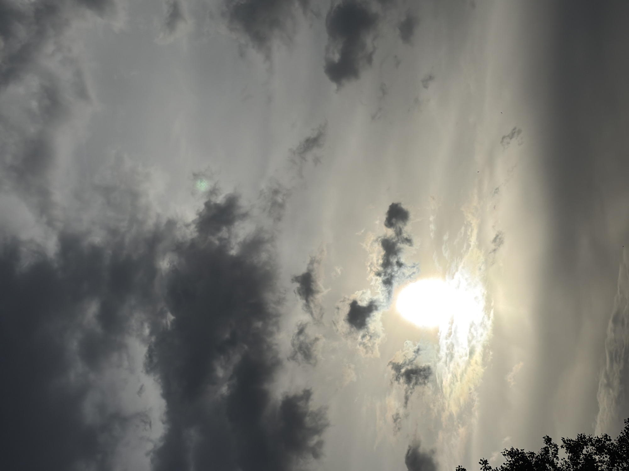 The image depicts a sky with heavy, dark clouds covering a significant portion of the frame. The sun is visible, partially obscured by thinner clouds, creating a bright, diffused light. The sky is gray with varying shades indicating different densities of cloud cover. In the bottom right corner of the frame, there is a glimpse of tree foliage. The location is not identifiable in the image. No people, specific objects, or actions are visible.