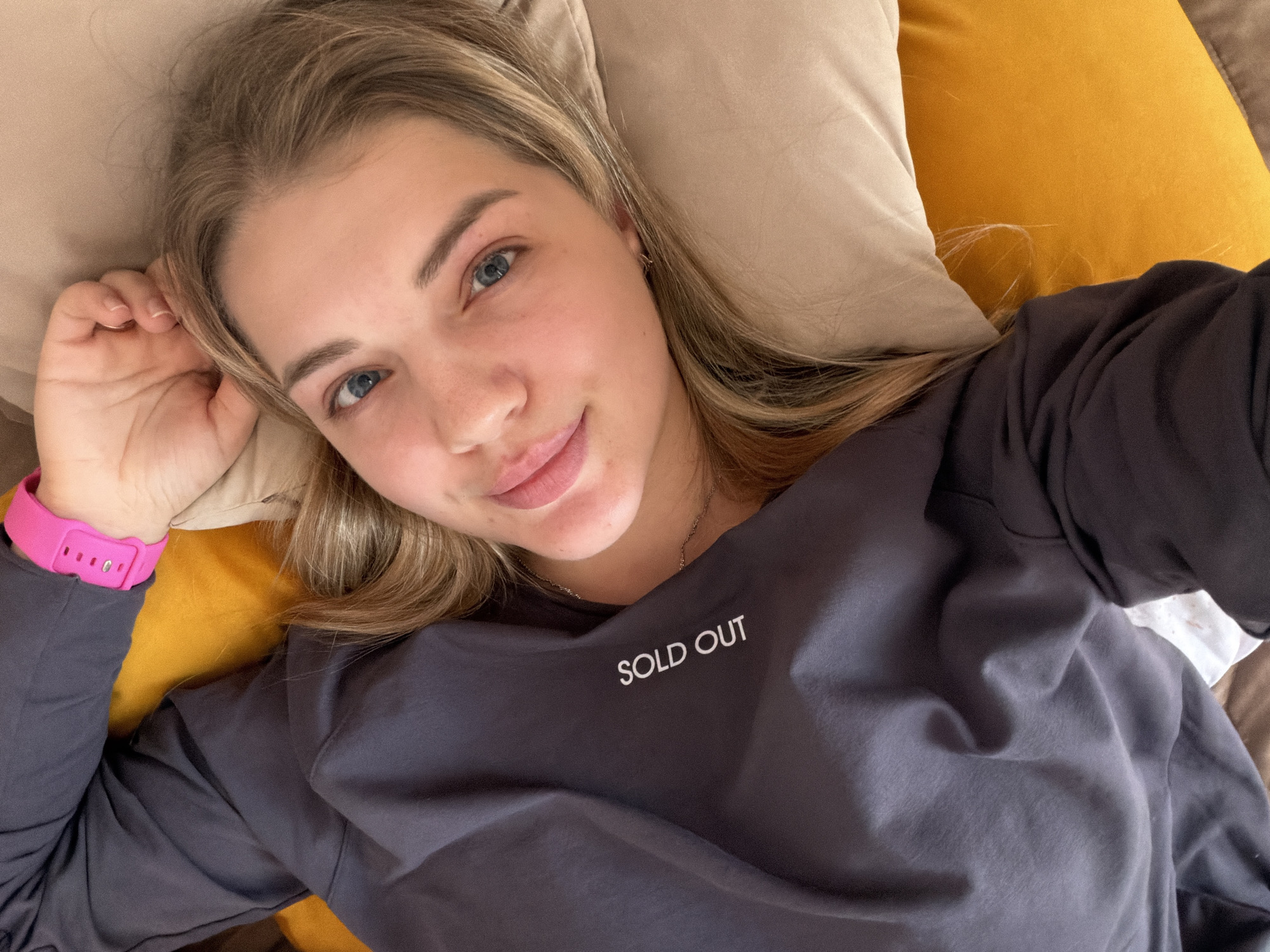 A young woman with blonde hair and blue eyes lies on an orange pillow and a beige pillow. She is wearing a long-sleeved gray shirt that reads "SOLD OUT." Her left wrist has a pink watch. The location is Большая Шильна, Russia.