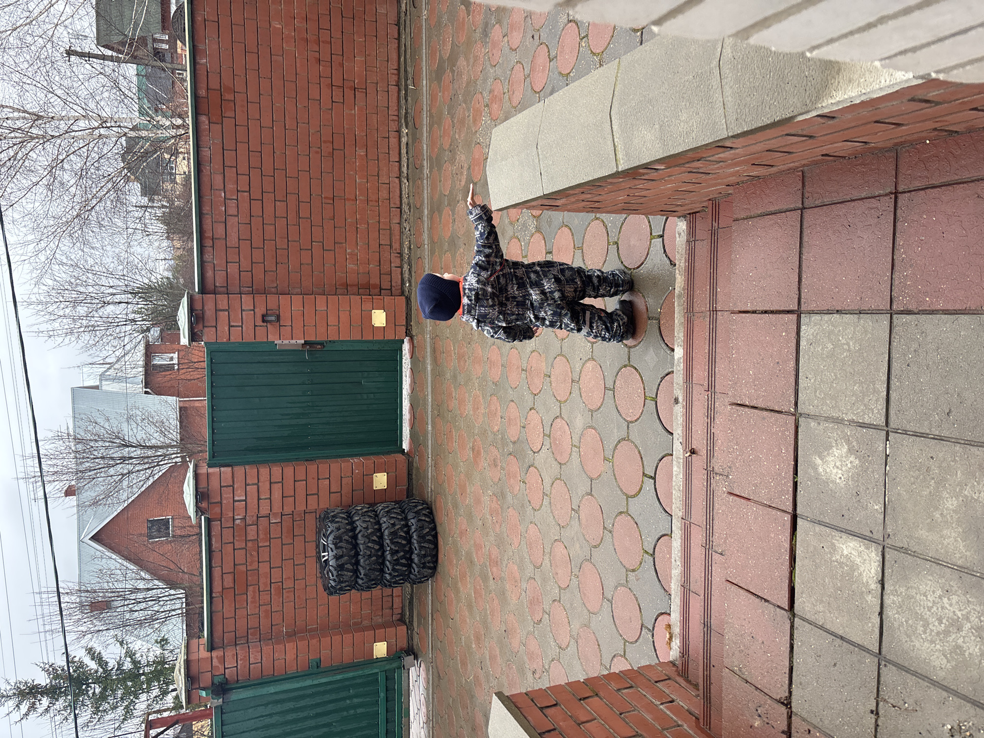 A child in a patterned jumpsuit and a dark blue hat stands on a tiled walkway. The walkway is made of patterned tiles with alternating grey and red circles. The child has their right arm raised. Behind the child, there is a brick wall with a green gate. Next to the gate, there are four stacked tires. In the background, there is a house and bare trees. The location is Shul'gino, Russia.