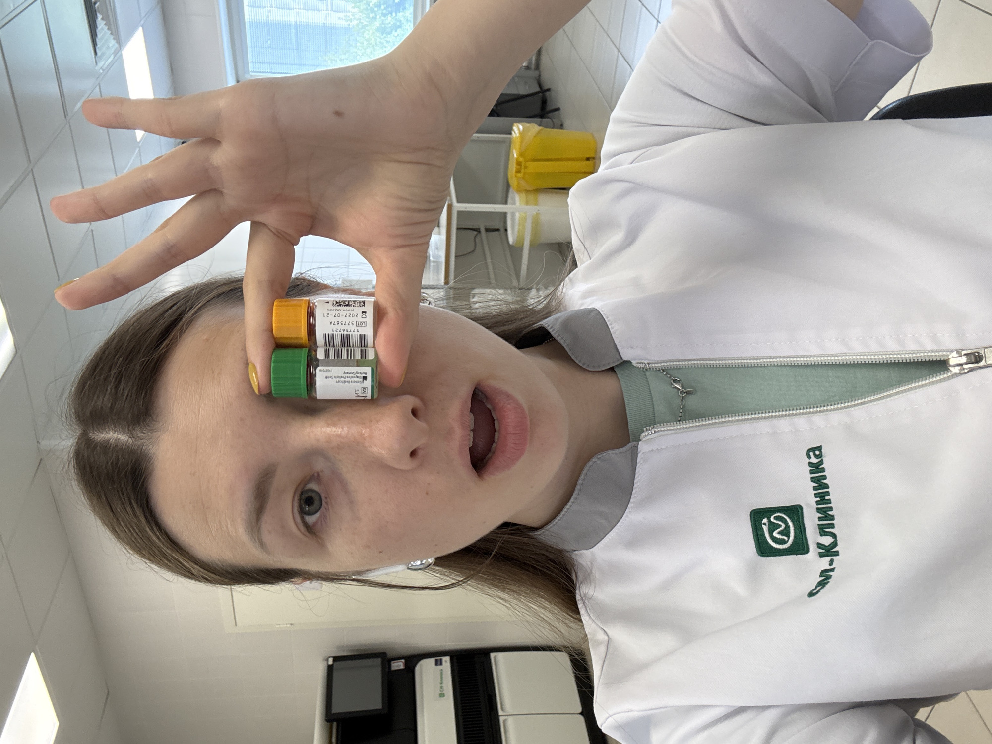 A woman with long brown hair and green eyes holds two small sample vials, one green-capped and one orange-capped, in front of her right eye. She is wearing a white lab coat with a green logo and Cyrillic text that reads "см-Клиника." The background appears to be a laboratory or medical facility with scientific equipment, including a large machine with a screen and a yellow biohazard bin. The overall impression is that of a medical professional in a work environment. The location is identified as Moscow, Russia.