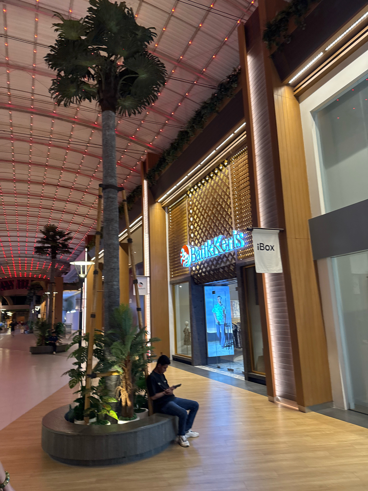 The image captures a view of an outdoor shopping arcade in Kuta, Indonesia, illuminated by ambient lighting. A tall, artificial palm tree stands prominently in the foreground, its trunk wrapped with dark bands. To the right of the palm tree, a shop storefront displays a brightly lit sign that reads "Batik Keris" in blue lettering, with a smaller banner hanging to its right that says "iBox". The shop's interior features a digital display showcasing a man in a patterned shirt. The flooring is made of light-colored wood planks, and a curved, concrete bench with potted plants surrounds the base of the palm tree. A young man, wearing a dark t-shirt and jeans, is seated on the bench, looking at his phone. Further down the arcade, other shops and people are visible, with a ceiling adorned with strings of red lights. The architecture suggests a modern, tropical-inspired mall.