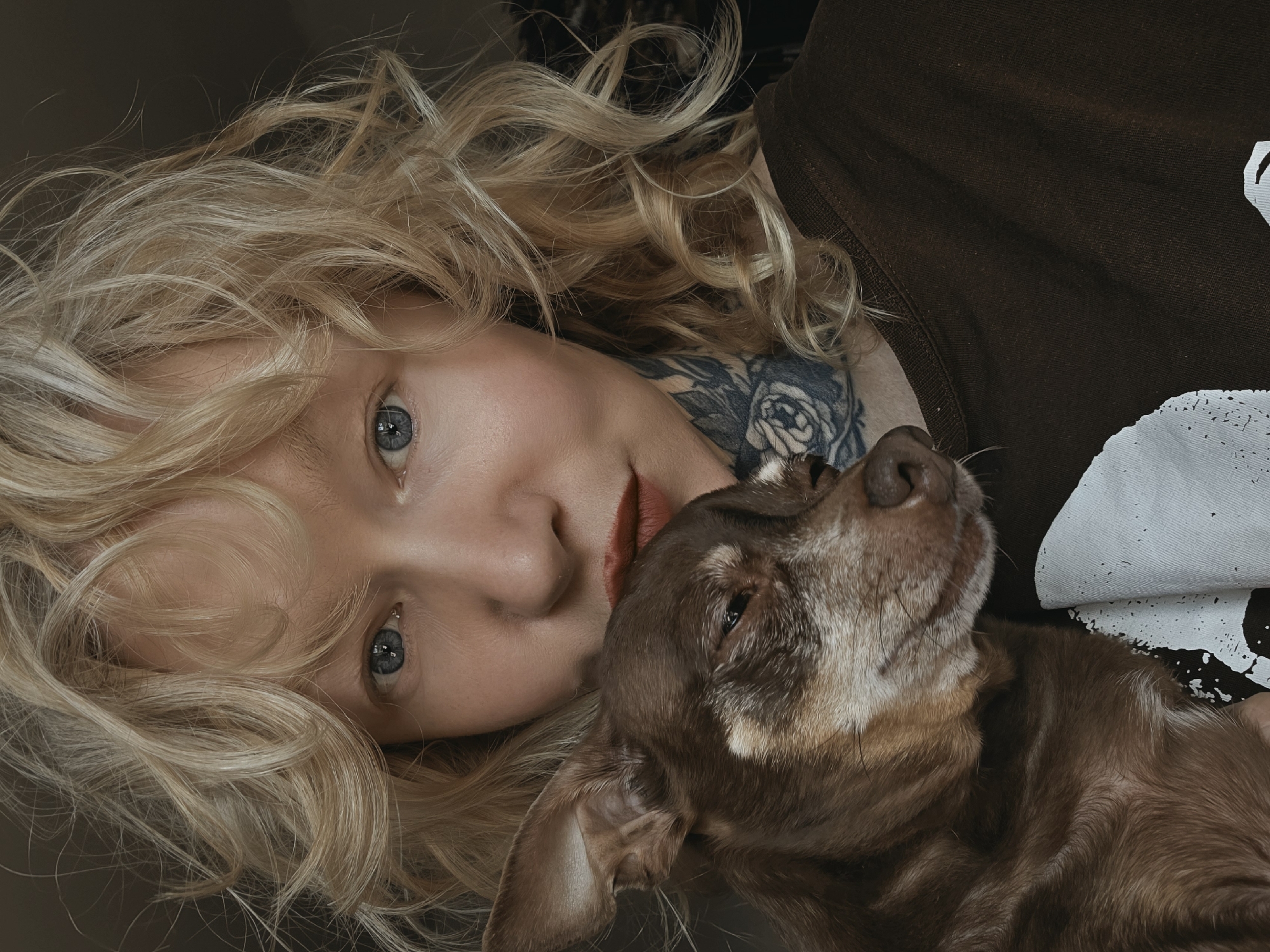 A woman with blonde, curly hair and blue eyes is holding a small, brown dog close to her face. She is wearing a dark brown t-shirt with a white graphic design on it and has tattoos on her neck. The dog has its eyes closed and appears to be resting against the woman. The lighting is dim, suggesting an indoor setting, possibly a home. There is no visible indication of the specific location within Saint Petersburg, Russia.