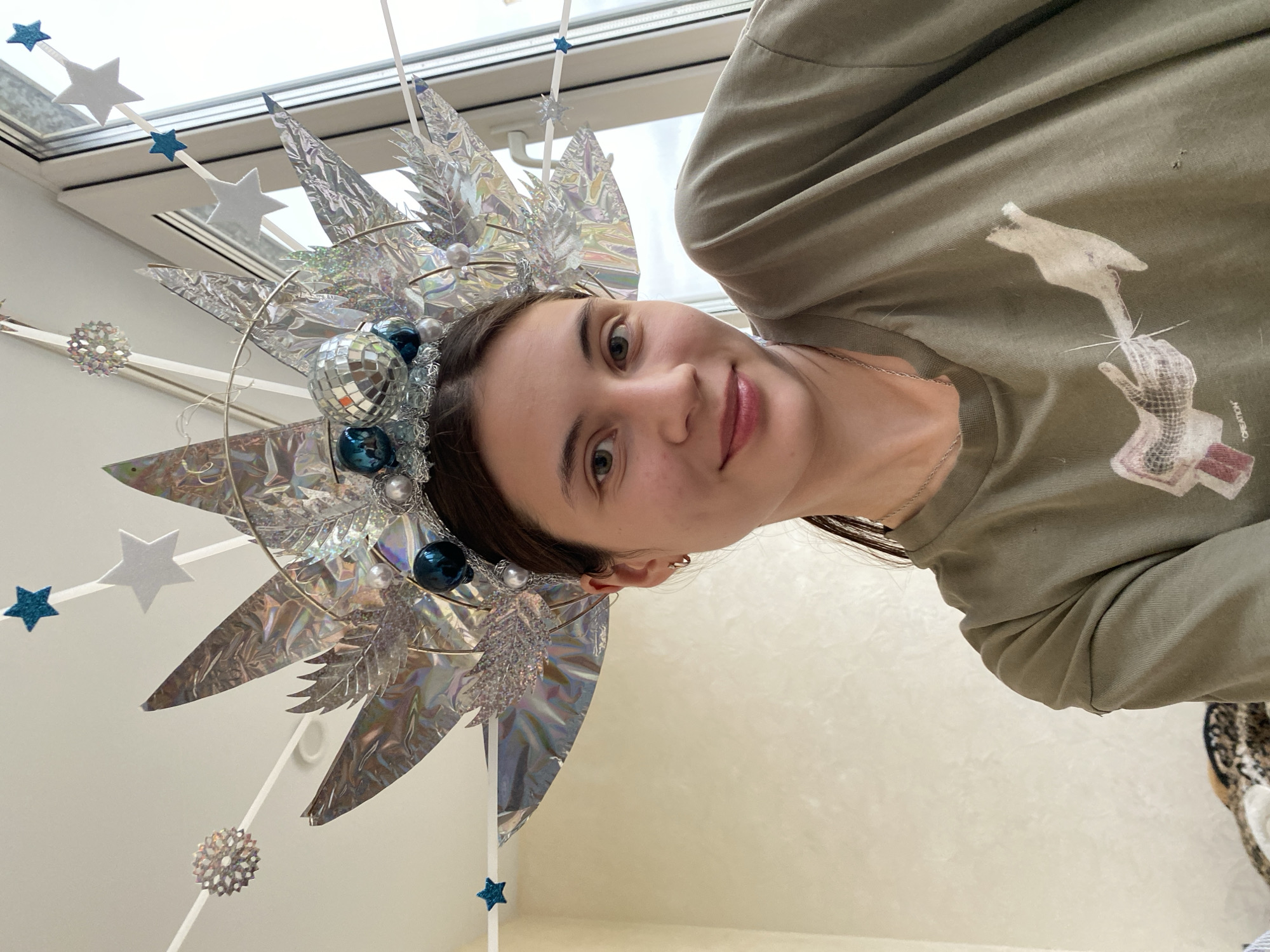 A young woman with brown hair is taking a selfie indoors, posing with a large, ornate, silver headdress that is decorated with blue baubles, mirrored spheres, and white and blue stars. She is wearing a green t-shirt with a graphic print on the front. The background shows a wall and a window with a view of a cloudy sky. The location is Votkinsk, Russia.