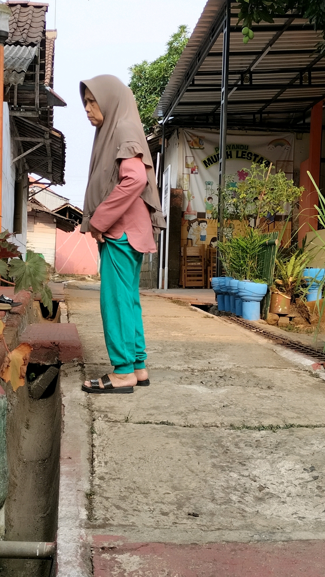 An elderly Indonesian woman wearing a taupe hijab and a salmon-colored long-sleeved shirt over teal sweatpants stands on a concrete path. She is facing left, with her head turned to look downwards. She wears black slide sandals. The scene appears to be in Subah, Indonesia, possibly a residential area or near a public building, with a covered walkway and a mural visible in the background. The ground is a worn concrete path with some vegetation growing through cracks. There is a drainage ditch to the left of the woman. Several blue planters are visible further down the path, along with potted plants. The overall atmosphere is casual and outdoors.