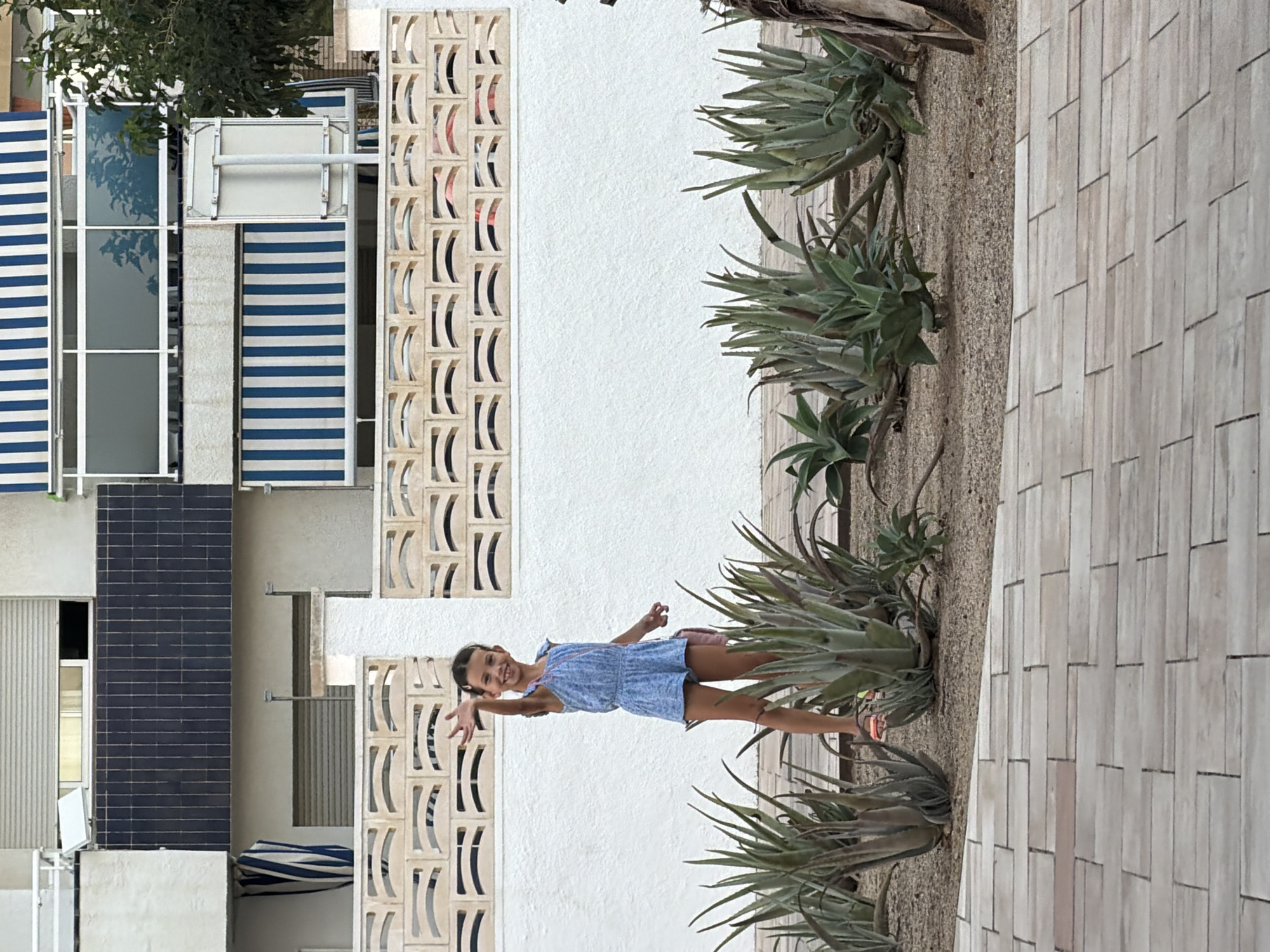 A young girl in a blue romper stands in front of a white wall and decorative concrete block screen. She is smiling and waving with her left hand. Behind her, a building with blue and white striped awnings and a dark tiled section is visible. The ground in front of the wall is covered with gravel and planted with aloe vera plants. The foreground shows a pathway made of rectangular beige tiles. The image appears to have been taken outdoors in a sunny environment. The location is identified as Mont-roig del Camp, Spain.