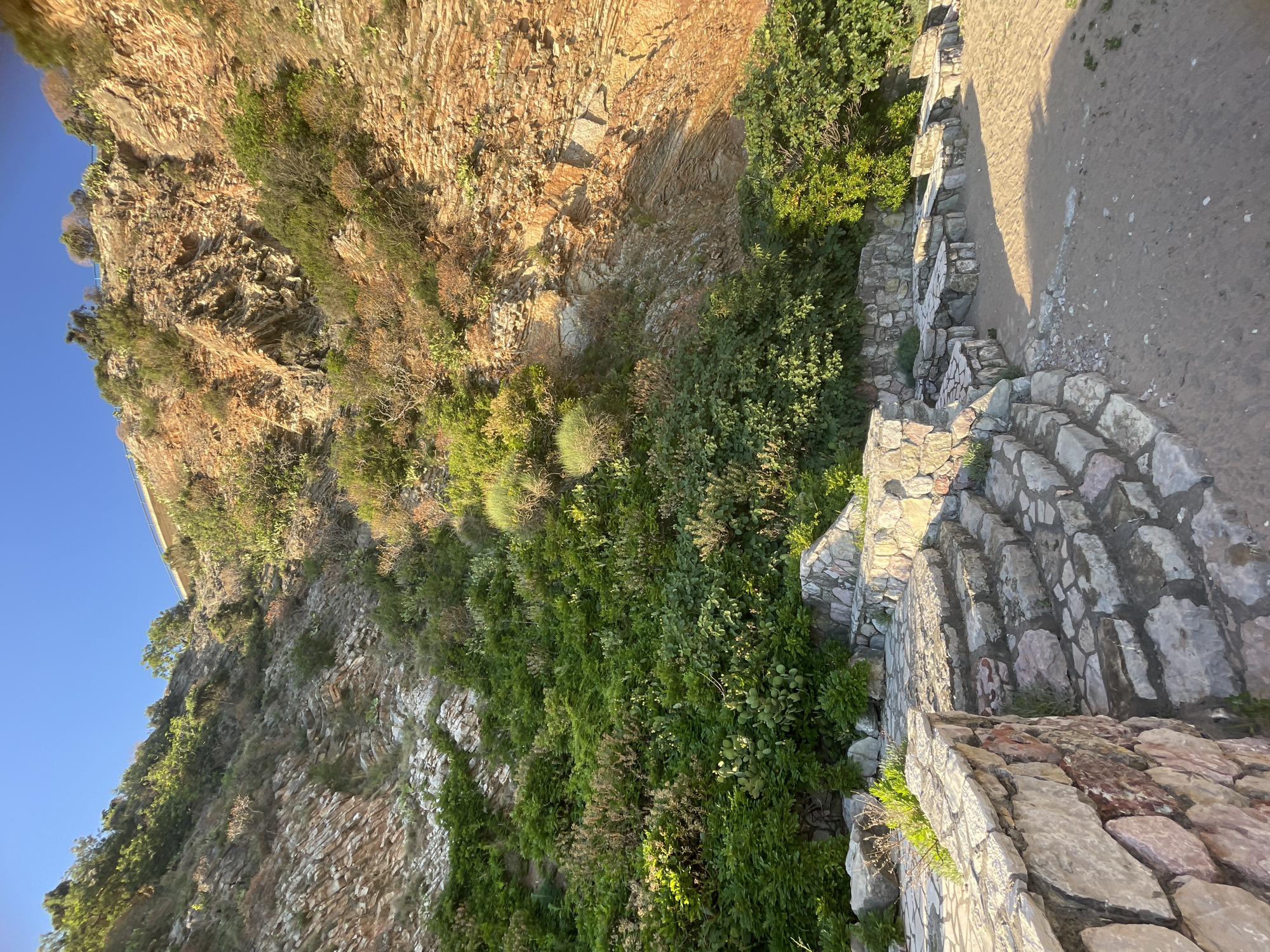 The image depicts a steep, rocky hillside in Rafailovići, Montenegro. The hillside is partially covered in dense green vegetation and reddish-brown exposed rock. A stone wall and steps made of irregular stone blocks run along the edge of the hillside, adjacent to a dirt path. The sky is visible in the upper left corner, appearing blue. The perspective is looking down from above.