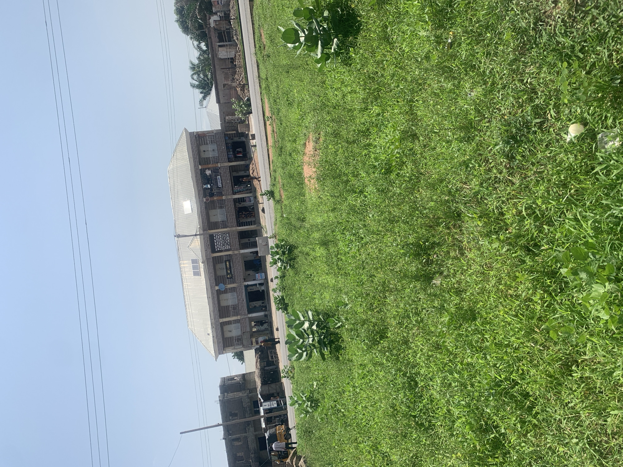 The image shows a street scene in Jalingo, Nigeria, with buildings lining one side and a grassy area on the other. The buildings appear to be commercial structures, with multiple stories and storefronts at ground level. One building has signage that reads "JEC LUXURY." People are visible walking on the street and near the buildings. The scene suggests it is daytime, with bright sunlight and a clear sky. The presence of power lines indicates an urban or semi-urban setting. The overall atmosphere appears to be that of a typical day in a Nigerian town.