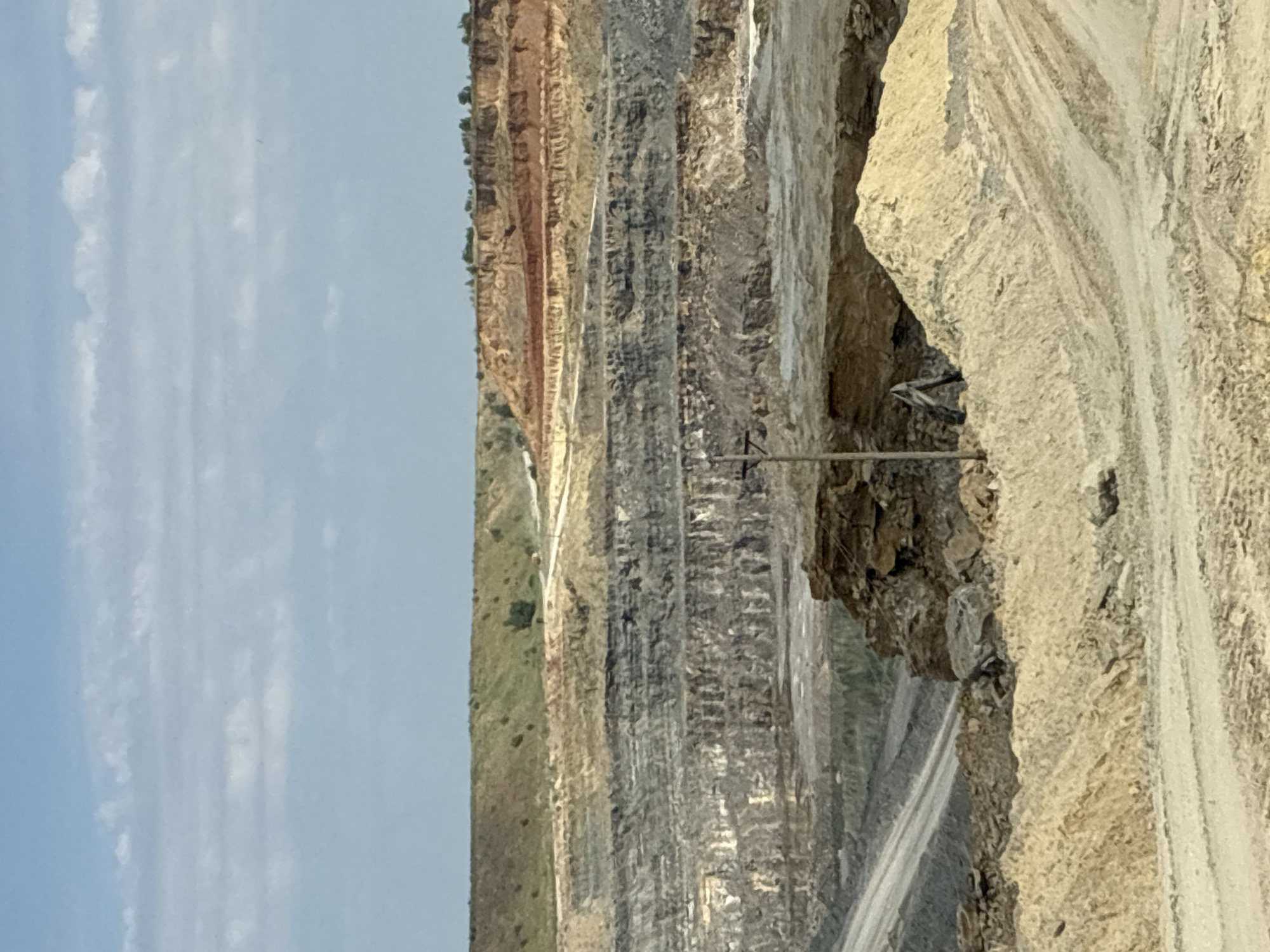 The image shows an aerial view of a large open-pit mine. The terrain is heavily excavated, with distinct layers of soil and rock formations visible. The sky is partly cloudy. In the foreground, there are piles of sand and dirt, with tire tracks suggesting recent vehicle activity. A single utility pole stands on the left side of the image, near the edge of the pit. The mine appears to be located in an arid or semi-arid region. The location is identified as Tatsinskaya, Russia.