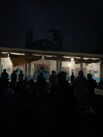 The image shows a gathering of people in front of what appears to be a building, likely a mosque, in the city of Kandahar, Afghanistan. The scene is dark, with minimal lighting, making it difficult to discern precise details. The building has a flat roof with a dome-like structure visible on top, as well as a minaret. A set of speakers is mounted above the dome. A series of columns and arches support the roof in front of the building. Many people stand and sit in front of the mosque, facing the building. The light makes it hard to discern facial features or clothing, but it seems they are gathered for some kind of event or prayer. The environment appears to be an open outdoor space.
