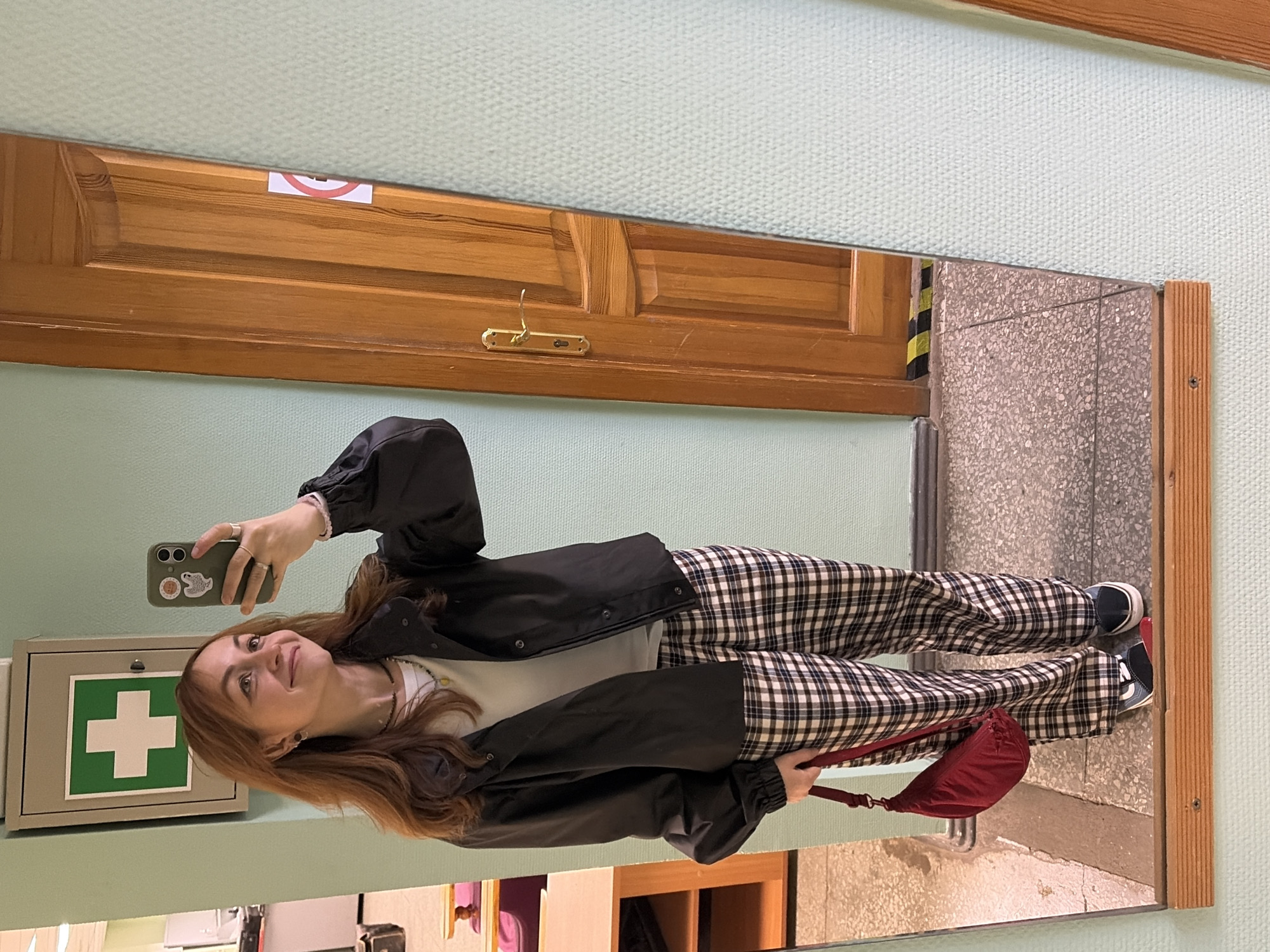 A young woman is captured taking a mirror selfie in an indoor setting, likely in Chaikovsky, Russia. The image is rotated 90 degrees clockwise. She has shoulder-length reddish-brown hair and smiles warmly, looking towards her dark-colored smartphone, which has a light-colored case featuring a snake-like design. She is wearing a black jacket over a light-colored top, black and white plaid pants, and dark sneakers. In her right hand, she holds a small, reddish-brown fabric shoulder bag.
The mirror reflects a room with a light green, textured wall. Visible in the reflection are a wooden door with horizontal panels, a brass-colored handle, and a coat hook. Above the door, a small sign with a red circle and a diagonal line, indicating prohibition, is partially seen. To the left in the reflection, a grey first aid kit with a prominent green cross symbol is mounted on the wall. The floor seen in the reflection is speckled light grey. A strip of black and yellow warning tape is also visible near the bottom right of the reflected door. The overall ambiance suggests a well-lit public or institutional building. The woman appears happy and relaxed.