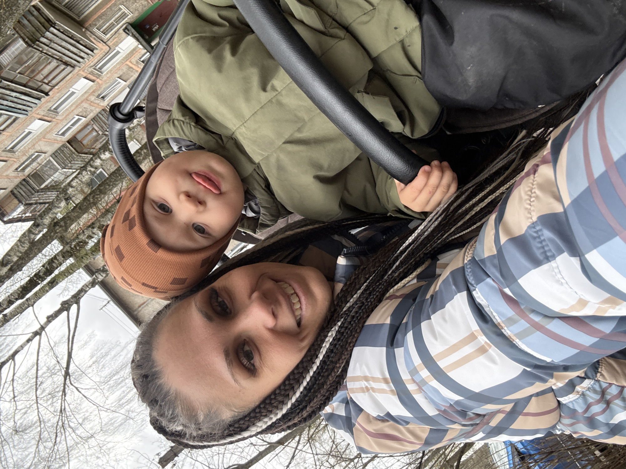 A woman and a child are in a stroller. The child is wearing a brown beanie and an olive-green jacket. The woman is wearing a plaid jacket and has braids. The location appears to be outside, possibly in a park or residential area, with buildings and bare trees visible in the background. The photo was taken in Rusanovo, Russia.