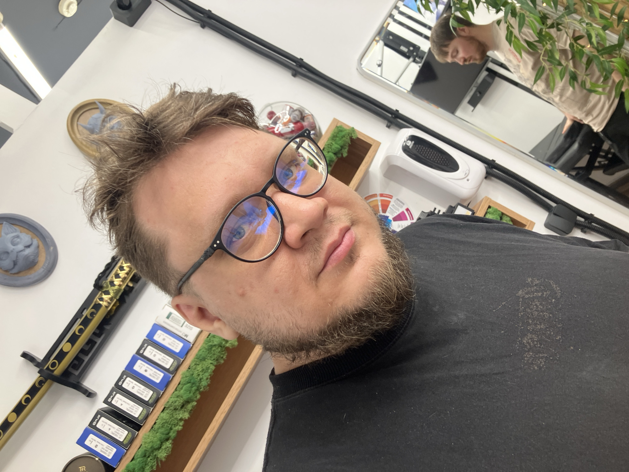 A man with a light brown beard and messy brown hair, wearing dark-framed glasses, captures a selfie. He looks directly at the camera with a neutral expression, his blue eyes visible through the lenses, reflecting indoor light sources. He is wearing a black crew-neck sweatshirt or t-shirt that appears slightly worn or dusty, with a faded pattern on the chest that resembles text but is illegible.
The setting appears to be an indoor space, possibly a personal studio, shop, or salon, located in Chaikovsky, Russia. The background is brightly lit, primarily with white walls. To the man's left, a decorative golden and black sword or katana with crescent moon motifs is mounted on a black stand. Below it, a wooden shelf holds multiple small product boxes, mostly black and blue, with white labels displaying text like "PUFF," "SIZE 100/30," "M," and "L." Above the sword, a framed, light blue three-dimensional sculpture of what looks like a stylized animal head or mythical creature is mounted on the wall.
To the man's right, a large rectangular mirror reflects another person, likely the same man, wearing a light-colored top and black trousers, with green foliage partially obscuring the reflection. Below the mirror, a white wall-mounted device, possibly a hand dryer or air purifier, is visible. Next to it, a wooden box contains green moss-like decor. Black corrugated tubes run along the wall. A colorful circular chart, potentially a color wheel, is partially visible. A small round container with a "TR" logo is also seen on a shelf.