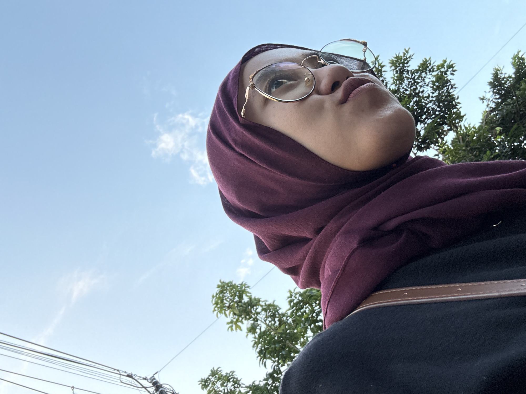 A low-angle shot depicts a person wearing glasses and a maroon headscarf, looking upwards. The background includes a blue sky with scattered clouds, as well as green trees and utility wires. The person is wearing a dark-colored shirt with a brown strap. The location appears to be Gadingrejo, Indonesia.