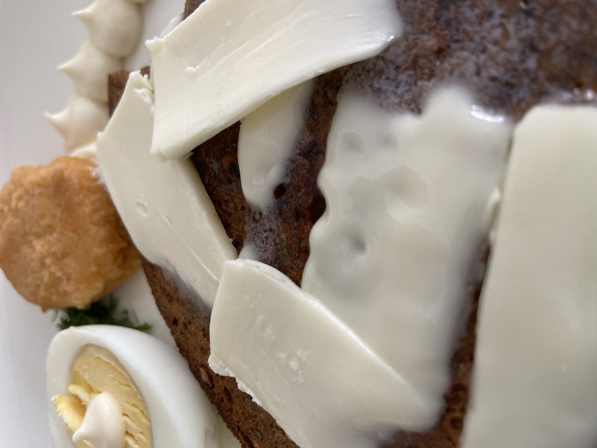 The image shows a close-up of a plate of food. The primary element is a slice of dark bread topped with several slices of a creamy white substance, likely cheese or butter. To the left, there is a boiled egg cut in half, with a dollop of white sauce on the yolk. A small, round, golden-brown item, possibly a pastry or croquette, is also present. A small sprig of dill is visible between the egg and the pastry. A row of decorative dollops of white cream or sauce is partially visible along the top left edge of the plate. The plate appears to be white. The composition is tightly cropped, providing a macro perspective on the food.
There is not enough information to conclude location.