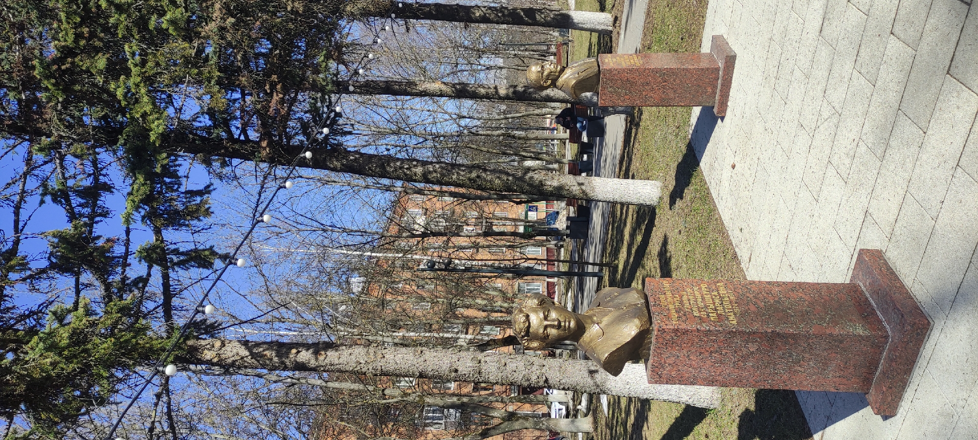 The image captures a park setting in Fryazino, Russia, featuring two bronze busts mounted on dark red granite pedestals. The busts appear to be portraits of individuals, positioned along a paved pathway. The environment includes trees, some with bare branches, others with green foliage. A building is visible in the background. Strands of lights are strung between the trees. A person sits on a park bench in the background. The ground is covered in grass and pavement.