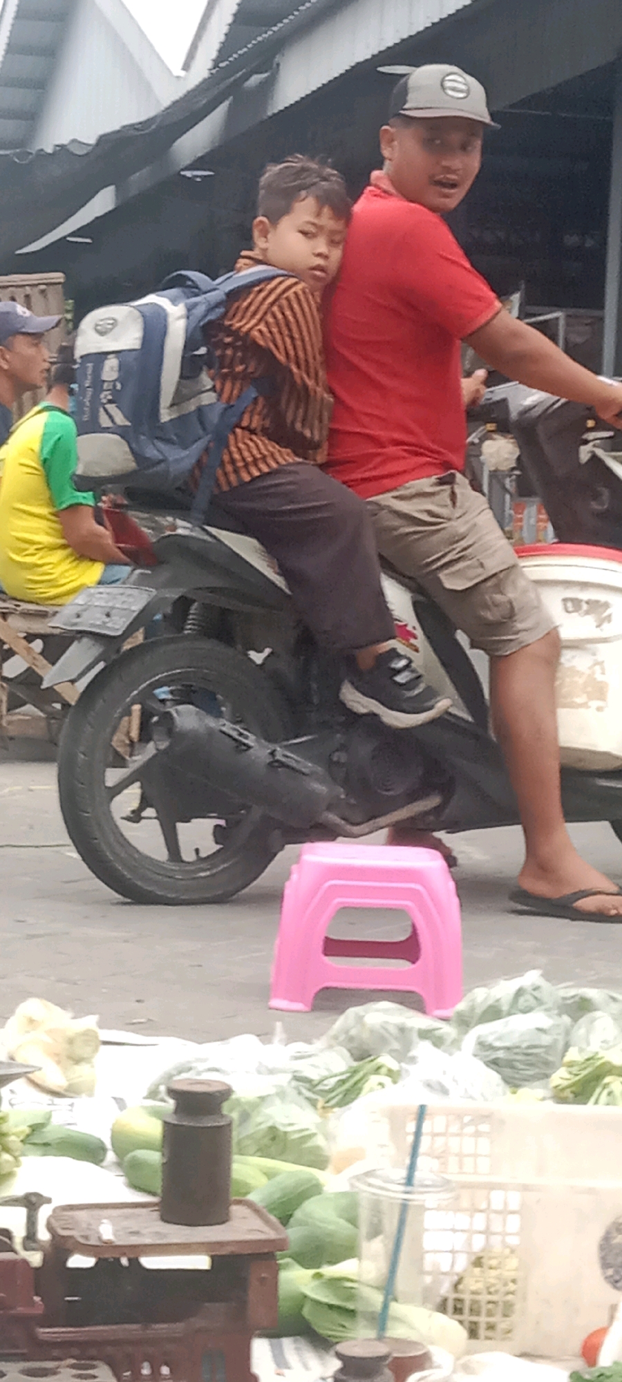 In Kras, Indonesia, a boy wearing a striped long-sleeved shirt and a blue backpack is riding on the back of a motorcycle, clinging to the driver who is wearing a red t-shirt and tan cargo shorts. The driver is looking towards the right, with his mouth open. Another man in a yellow shirt and green sleeves is seated on a wooden structure to the left of the motorcycle. The motorcycle is parked on a paved surface, with a pink plastic stool in the foreground. Behind the motorcycle, there are various vegetables and produce spread out, some of which are bagged in plastic. A vintage scale with a weight is visible on the left side of the frame. The scene appears to be a marketplace or street vendor area.
