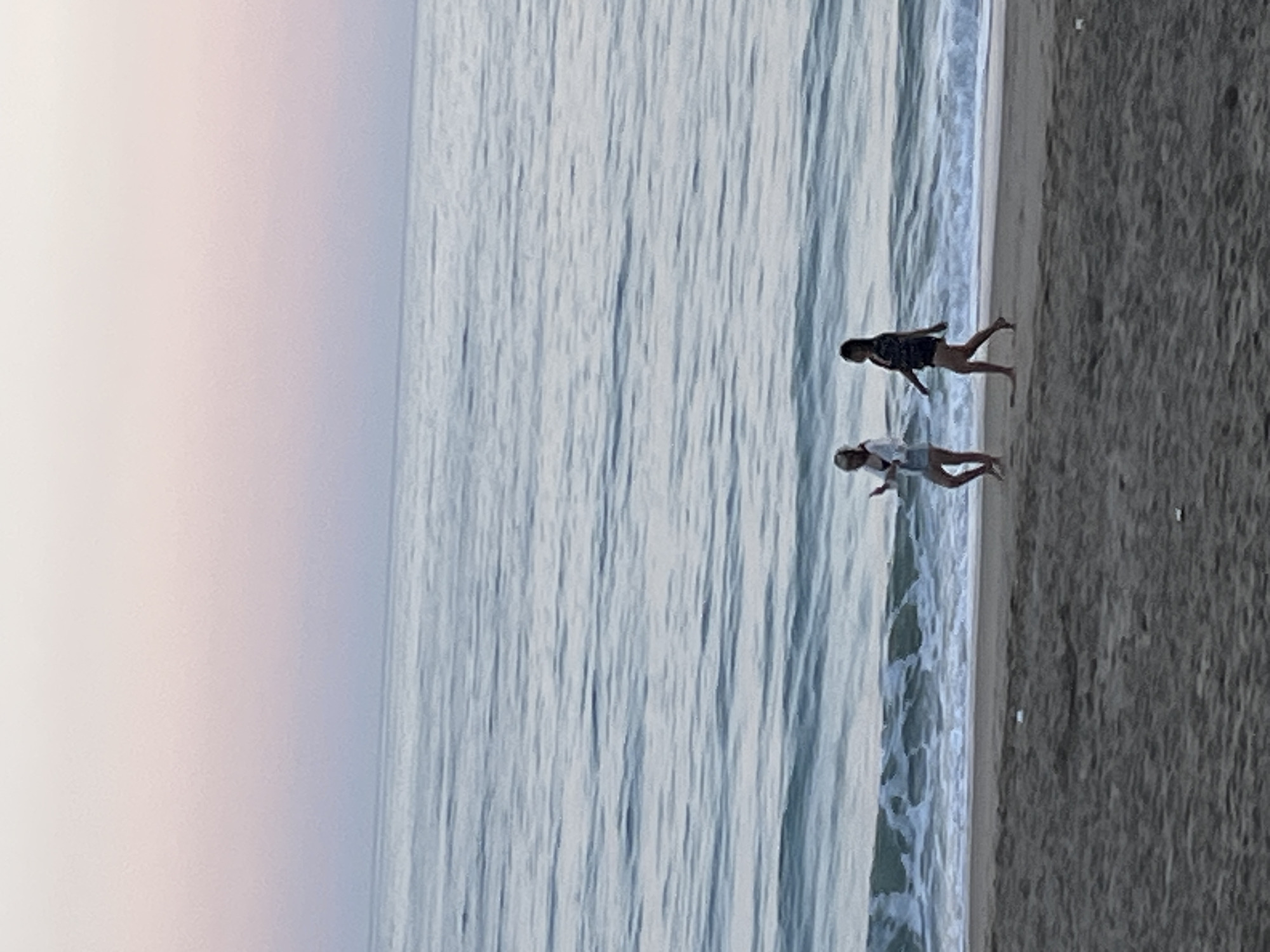 Two individuals are observed on a beach adjacent to the sea. The location is presumed to be in Alanya, Turkey. One individual is wearing dark clothing, while the other is in lighter apparel. They are positioned near the water's edge. The environment includes the sea, sand, and sky. The sky appears to be overcast.