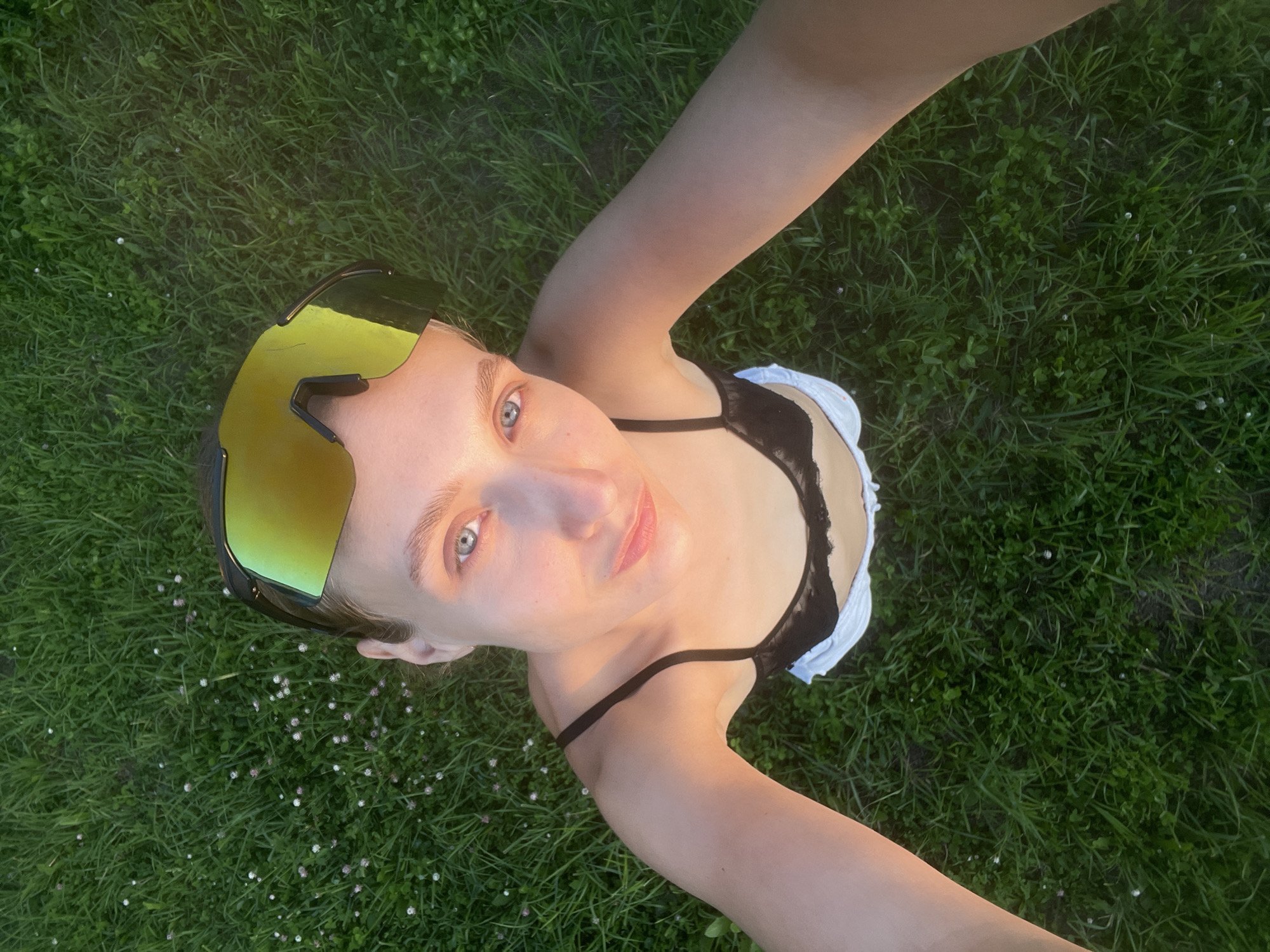 A high-angle, close-up, selfie-style shot shows a young person with blonde hair wearing reflective yellow sunglasses and a black bralette over white shorts, lying on a green grassy field. The person's face is tilted towards the camera, with their eyes open and looking upwards. The grass is lush and green, with small white and pink flowers interspersed throughout. The lighting suggests it is either early morning or late evening, casting a warm glow on the person's skin. The background is entirely covered by the grassy field. The image does not contain any information about the city of Подшивалово, Russia.