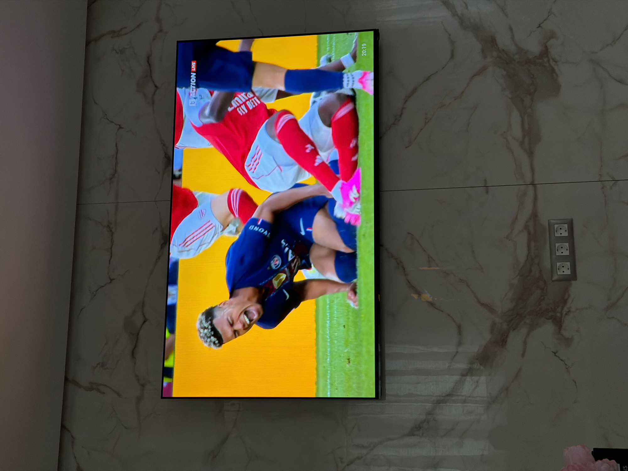 The image shows a wall-mounted television in a room. The wall is covered in a marbled pattern, possibly made of tile or stone. To the right of the television, there is a power outlet. The television screen displays a soccer game in progress, with several players visible in mid-action. A player in a blue uniform appears to be falling or being tackled. The background of the game shows a yellow field. Text on the screen shows "ACTION LIVE" and "20:19", which appear to be related to the game broadcast. The location of the image is Asenovgrad, Bulgaria.
