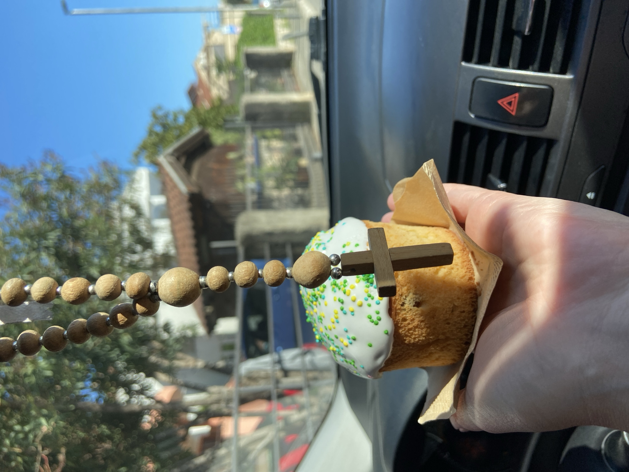 A hand holds a slice of Easter cake adorned with white frosting, green and yellow sprinkles, and a small wooden cross. A beaded rosary hangs in the foreground. The scene appears to be inside a vehicle, with a dashboard and emergency light button visible. Outside the window, there is a blurred view of buildings, trees, and a blue sky, suggesting an urban environment. Based on the building style and general layout, this photo was taken in Budva, Montenegro.