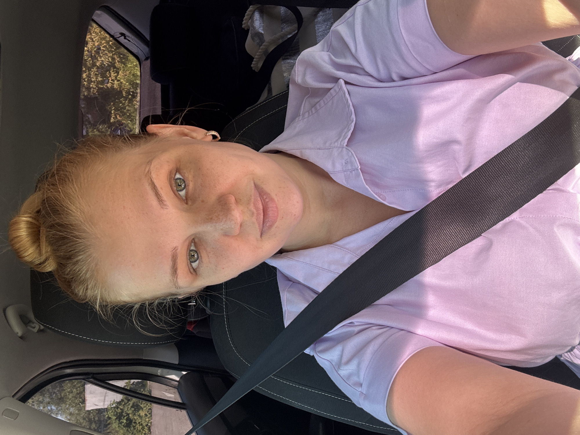 A blonde woman with her hair in a bun is sitting in the driver's seat of a car. She is wearing a light purple collared shirt and has a seatbelt fastened across her chest. She is looking directly at the camera with a neutral expression. The interior of the car is visible, including the headrest and the back seat. Sunlight is streaming into the car, casting shadows on her face and shirt. The background outside the car windows shows trees and foliage, suggesting an outdoor, possibly rural or suburban setting. The location is identified as Rusanovo, Russia.