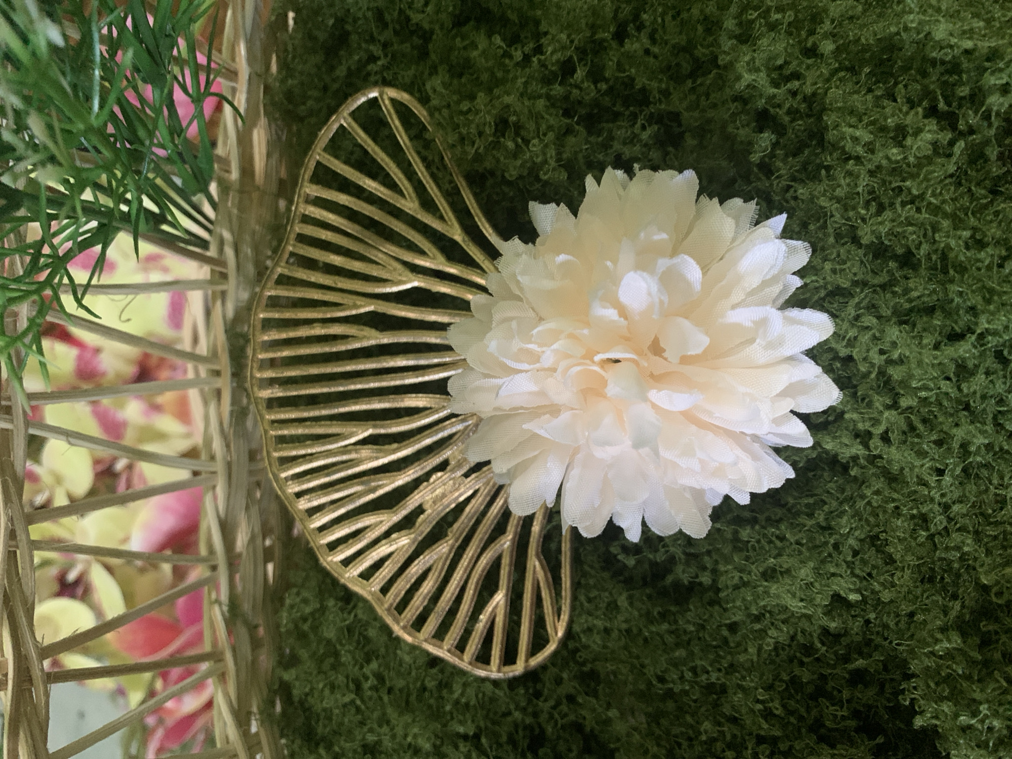 The image shows a close-up view of a decorative arrangement on a green mossy surface. In the foreground, a pale peach artificial flower with many delicate petals is centered. Behind the flower, a golden metallic decorative element, shaped like a fan or leaf with radiating lines, is positioned upright. The background reveals a wicker basket with a patterned fabric inside, featuring pink and yellow colors, and some green artificial foliage extending from the top. The setting appears to be an indoor decorative display, possibly for an event or as part of a larger arrangement. There is no discernible connection to Kuala Pembuang, Indonesia.