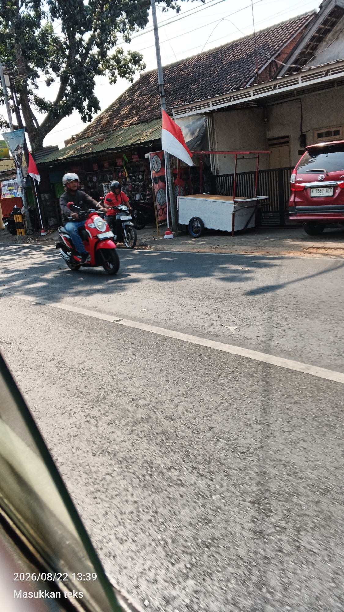 In Malang, Indonesia, a street scene unfolds with a red scooter carrying a person in a helmet and blue jeans. Behind them, another scooter is ridden by a person wearing a red shirt and helmet. The background features a building with a tiled roof, an Indonesian flag flying, and a white food cart. A red car with a license plate reading "N 1567 ACR" is parked on the right side of the street. The road is asphalt, with a white line marking the lane. The timestamp "2026/08/22 13:39" and the text "Masukkan teks" are visible in the lower left corner of the image, indicating it was taken on August 22, 2026, at 1:39 PM, and the user is prompted to enter text.