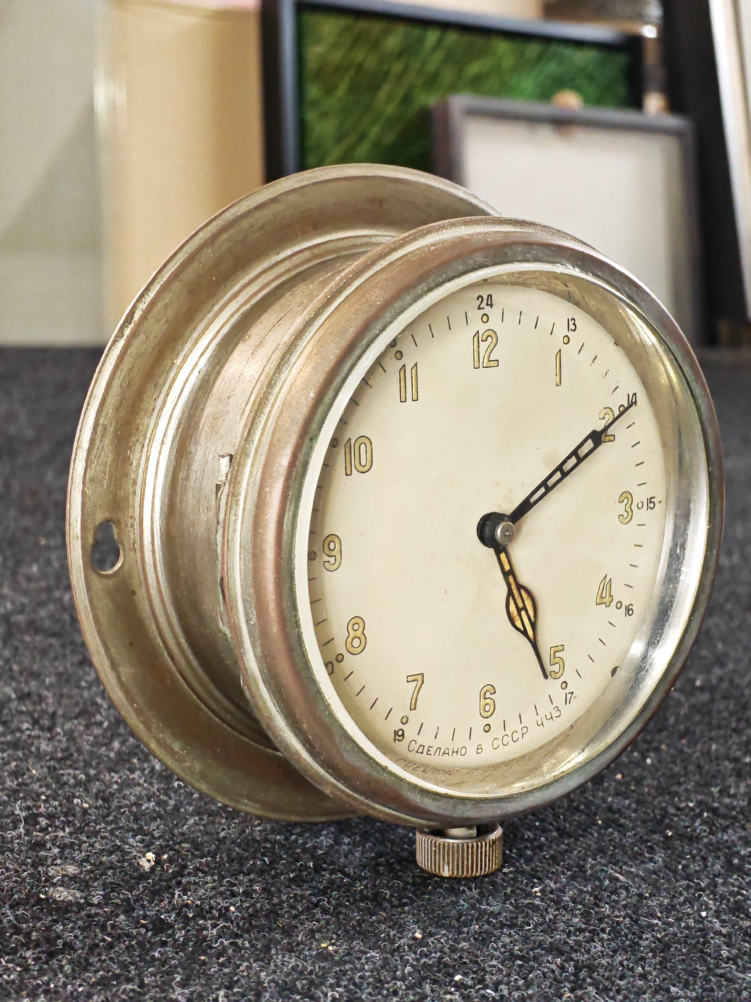 This is a close-up, slightly angled shot of an antique, circular clock with a metallic casing. The clock face is off-white with golden Arabic numerals marking the hours, from 1 to 12, as well as a 24-hour dial. There are two hands on the clock; the hour hand is shorter and points to the number 5, while the minute hand is longer and points to approximately the 14-minute mark. The casing appears to be made of brass or a similar metal and shows signs of age and wear, with a dull, brushed finish and some patina. A small winding knob is visible at the bottom of the clock.
The background is out of focus, showing what appears to be an indoor setting with objects that could be furniture or frames. The lighting suggests daytime. Text on the clock face, in Cyrillic, reads "СДЕЛАНО В СССР", which translates to "Made in USSR". There are also some numbers and symbols that are difficult to decipher completely due to the resolution and angle. The clock is resting on a dark, textured surface, possibly fabric or carpet.