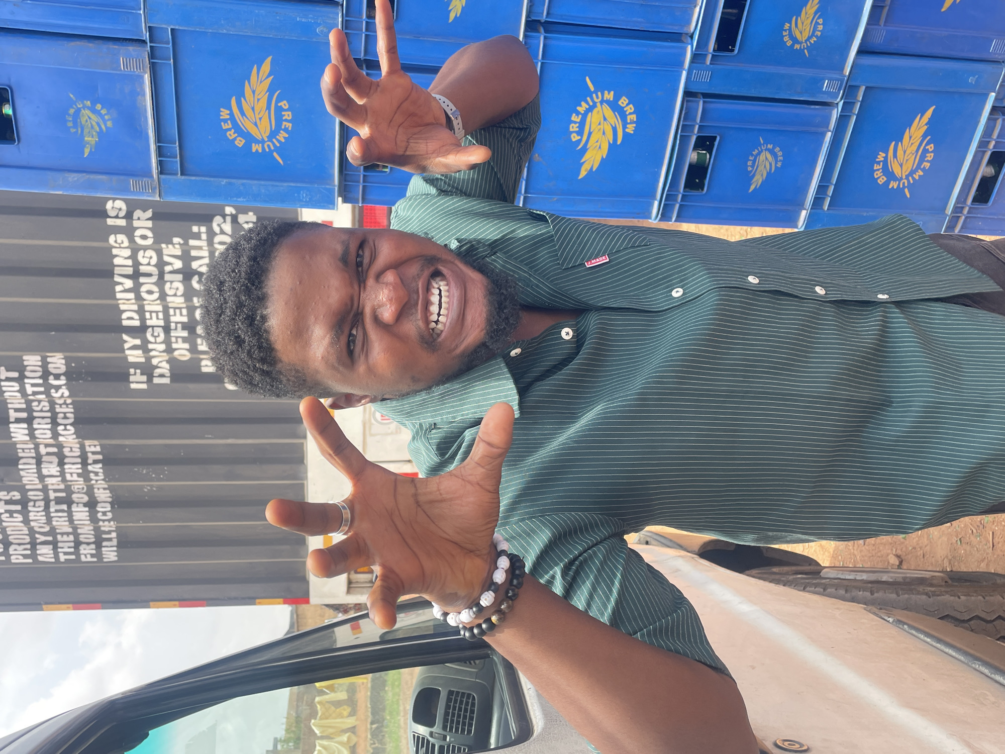 A man with dark skin, short black hair, and a beard is posing with claw-like hands in front of a stack of blue crates with the words "PREMIUM BREW" in yellow text and a logo resembling a wheat stalk, possibly in Gura Top, Nigeria. He wears a dark green pinstriped shirt and a ring on one hand, along with two bracelets. Behind him is a black container with printed text, including a warning about unauthorized cargo. A car is partially visible in the foreground.