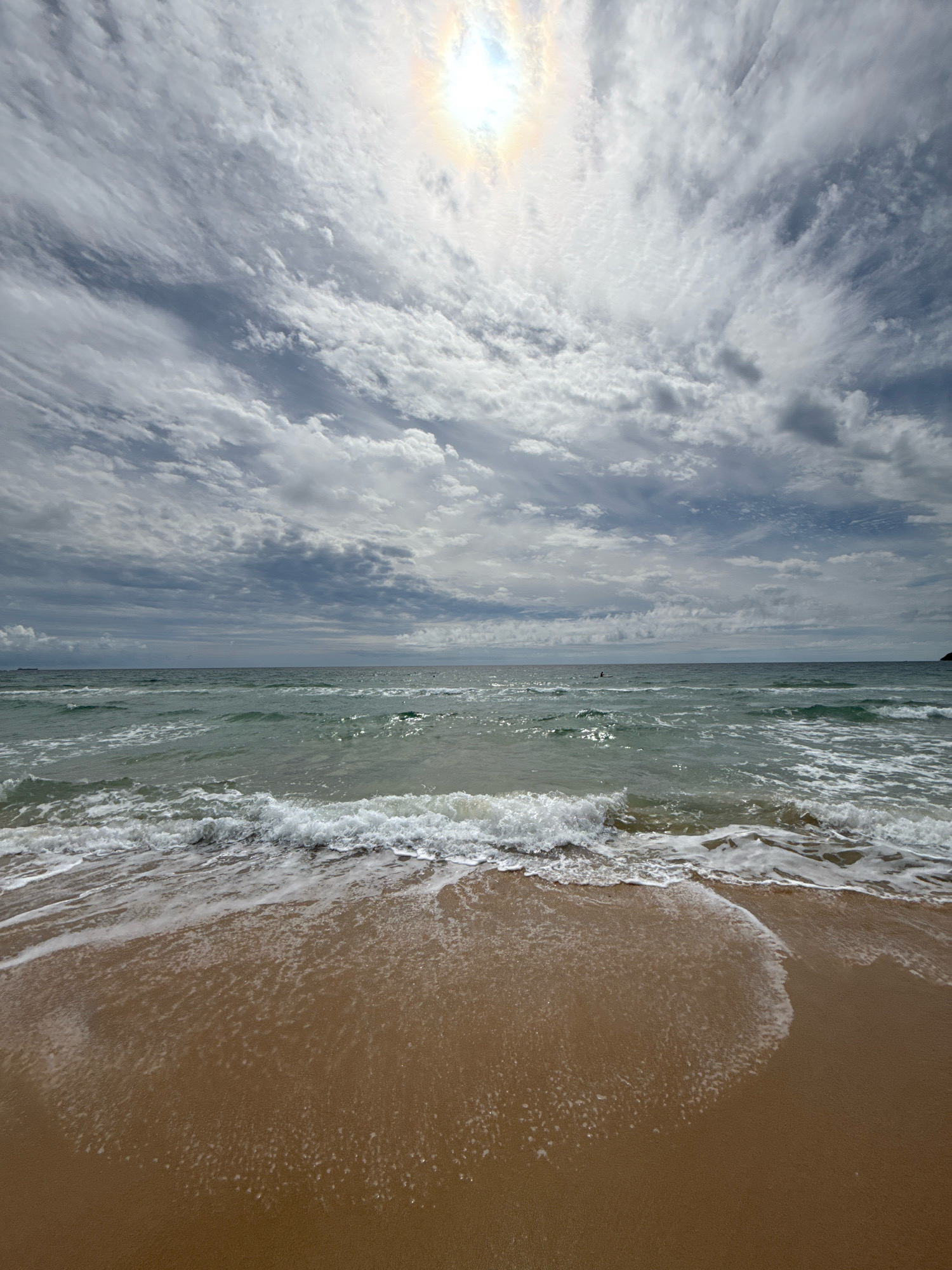 A wide-angle, eye-level shot captures a tranquil beach scene under a dramatic, cloud-filled sky. The sun, partially obscured by clouds, creates a bright, diffused glow at the top center of the frame, with hints of a rainbow visible. Wispy, elongated clouds stretch across the sky, with darker, more textured clouds interspersed throughout. The ocean, a deep green, exhibits gentle waves that are breaking near the shore, creating a foamy white surf that recedes onto the light brown sand. The sand occupies the lower portion of the image, with wet sand reflecting the light and dry sand appearing a warmer, more golden hue. In the distance, a faint line of the horizon separates the sea from the sky. Far out at sea, a tiny dark shape, possibly a distant boat or person, is visible. The overall mood is one of peaceful, natural beauty, with the dynamic sky contrasting with the calm sea and shore. The image does not depict the city of Anna, Russia; it is a generic beach landscape.