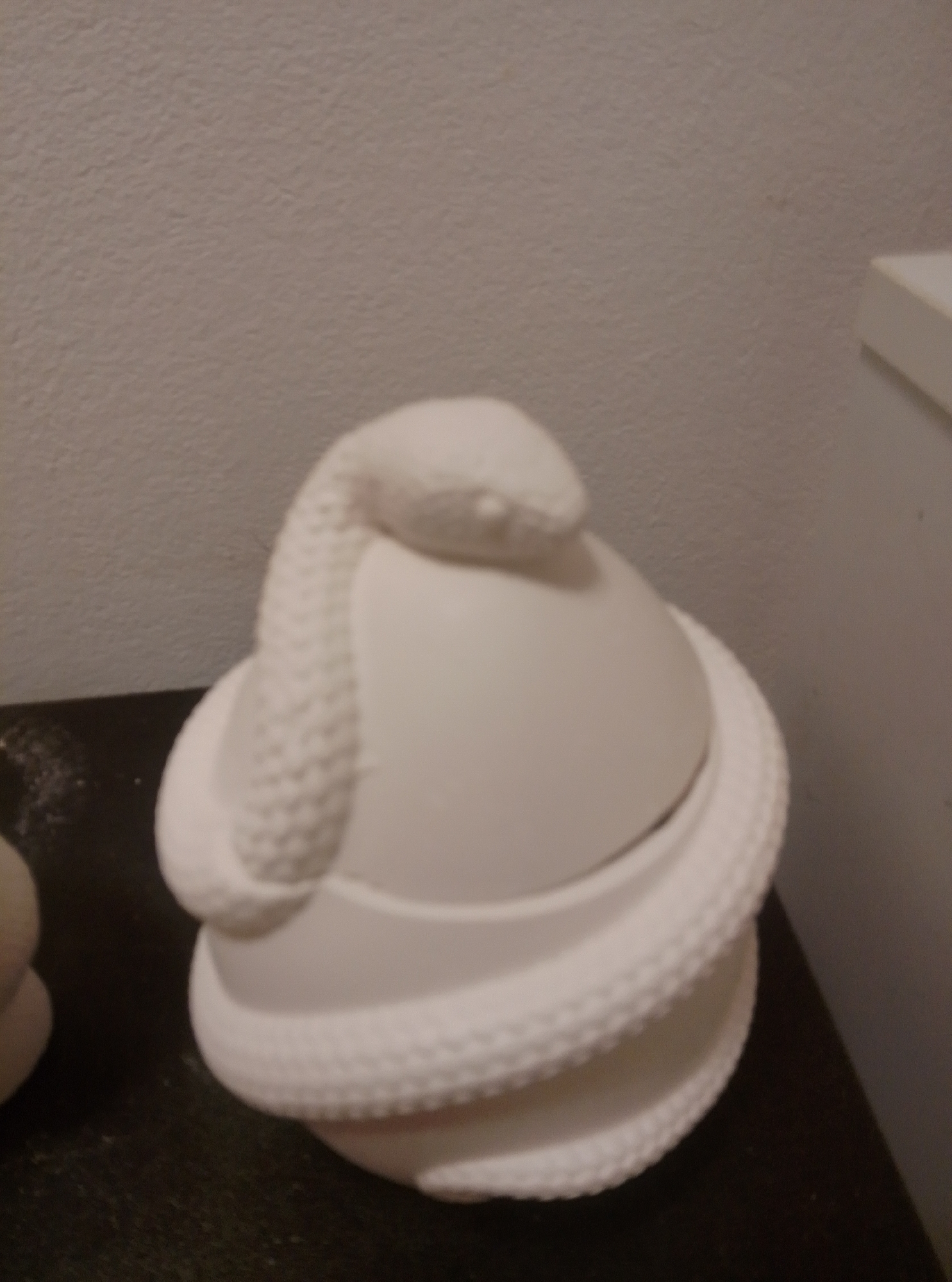 A white ceramic owl figurine with an attached coiled snake is displayed on a dark surface against a light-colored textured wall. The owl's head is visible at the top, with the snake's body wrapping around it and down the side. The snake's texture is detailed, resembling scales or a woven pattern. The bottom of the owl appears to be a jar or container, with a textured rim. The lighting is dim, casting soft shadows. The owl figurine is likely an unfinished ceramic piece. The scene is likely within an indoor setting, such as a home or studio, and no specific location in Wedel, Germany, can be identified from the visual information provided.