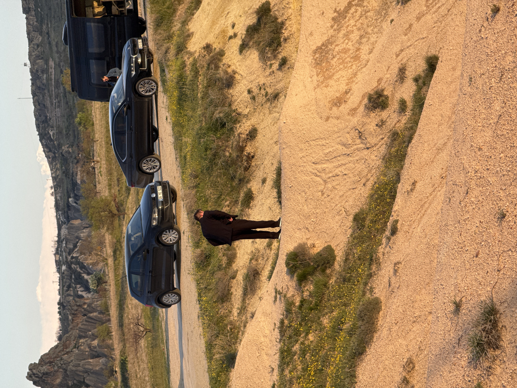 An outdoor landscape shot, rotated 90 degrees clockwise, depicts a man and several parked vehicles in an arid, rocky environment consistent with Göreme, Turkey.
The primary subject is a man dressed in all black, including a jacket and trousers, with white-soled sneakers. He stands on a light brown, sandy path or road, slightly elevated from a vegetated ditch to his left. His posture suggests he is observing the surroundings.
Along the adjacent dirt road to the left, three dark-colored vehicles are parked in a line. Closest to the foreground is a dark hatchback or small sedan (possibly a Volkswagen, indicated by a faint logo), with a visible license plate "309 TE 532". Behind it is a black sedan, likely a Skoda, with the license plate "34 BD 9527". Further back is a large, black van, possibly a Mercedes-Benz Sprinter, also showing the license plate "34 BD 9527".
The setting is characterized by eroded, sandy-colored hills and slopes, with sparse green vegetation, including small bushes and patches of yellow wildflowers. In the far background, distinct, layered rocky formations or mountains rise under a clear sky, typical of the Cappadocia region.
The time of day appears to be during the golden hour, either early morning or late afternoon, as indicated by the warm, soft light casting elongated shadows from the man and the vehicles onto the sandy ground. The overall mood is calm and serene.