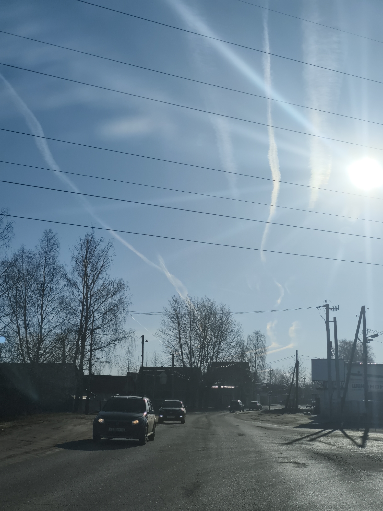 The image depicts a street scene in an unspecified city. The sky is blue with vapor trails and power lines running across it. The sun is shining brightly on the right side of the frame. Several cars are traveling on the road. Trees line the side of the road, most are bare of leaves. There is a building in the distance to the right, the signage is partially visible.