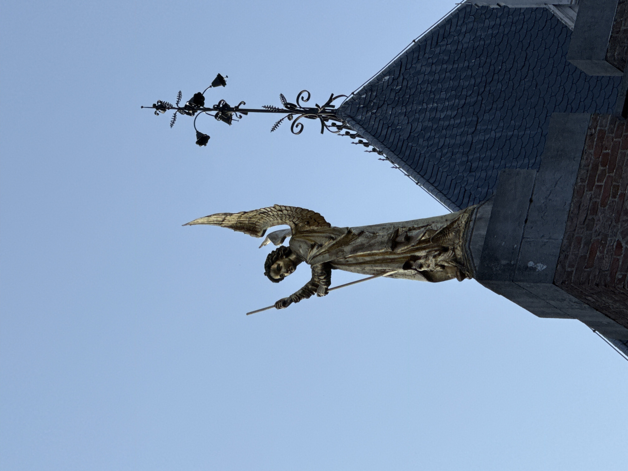 The image shows a golden statue of an angel atop a building, likely a church spire, in Bruges, Belgium. The angel is depicted with outstretched wings, holding a staff in its left hand. The statue appears to be made of bronze or gilded metal. Behind the angel, a dark, steep-pitched roof is visible, characteristic of gothic architecture. A decorative weather vane with bell-shaped elements is also present on the roofline. The sky is a clear, pale blue, indicating daylight. The building's base is constructed of brick. The statue is positioned on a stone plinth.