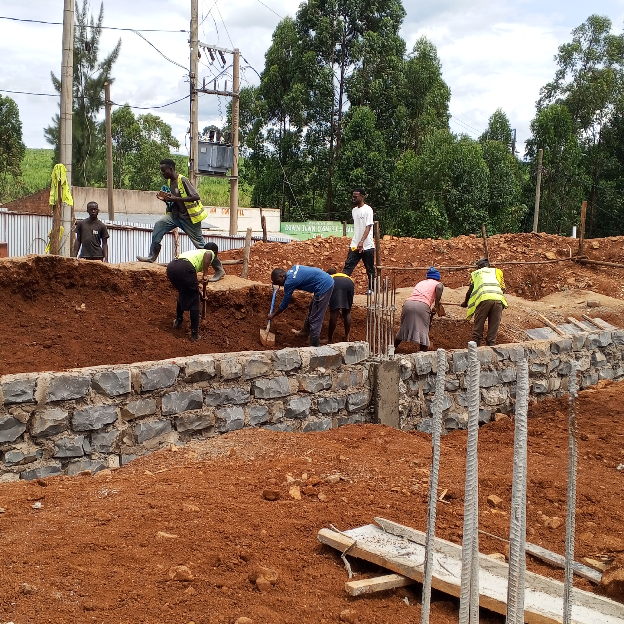 The image shows a construction site in Borabu Chitago ward, Kenya. Several people are working on the site, using shovels to move dirt. A stone wall has been partially constructed, with concrete pillars and reinforcing bars visible. Construction materials are scattered on the ground. In the background, there are buildings, trees, and utility poles. The sky is overcast.