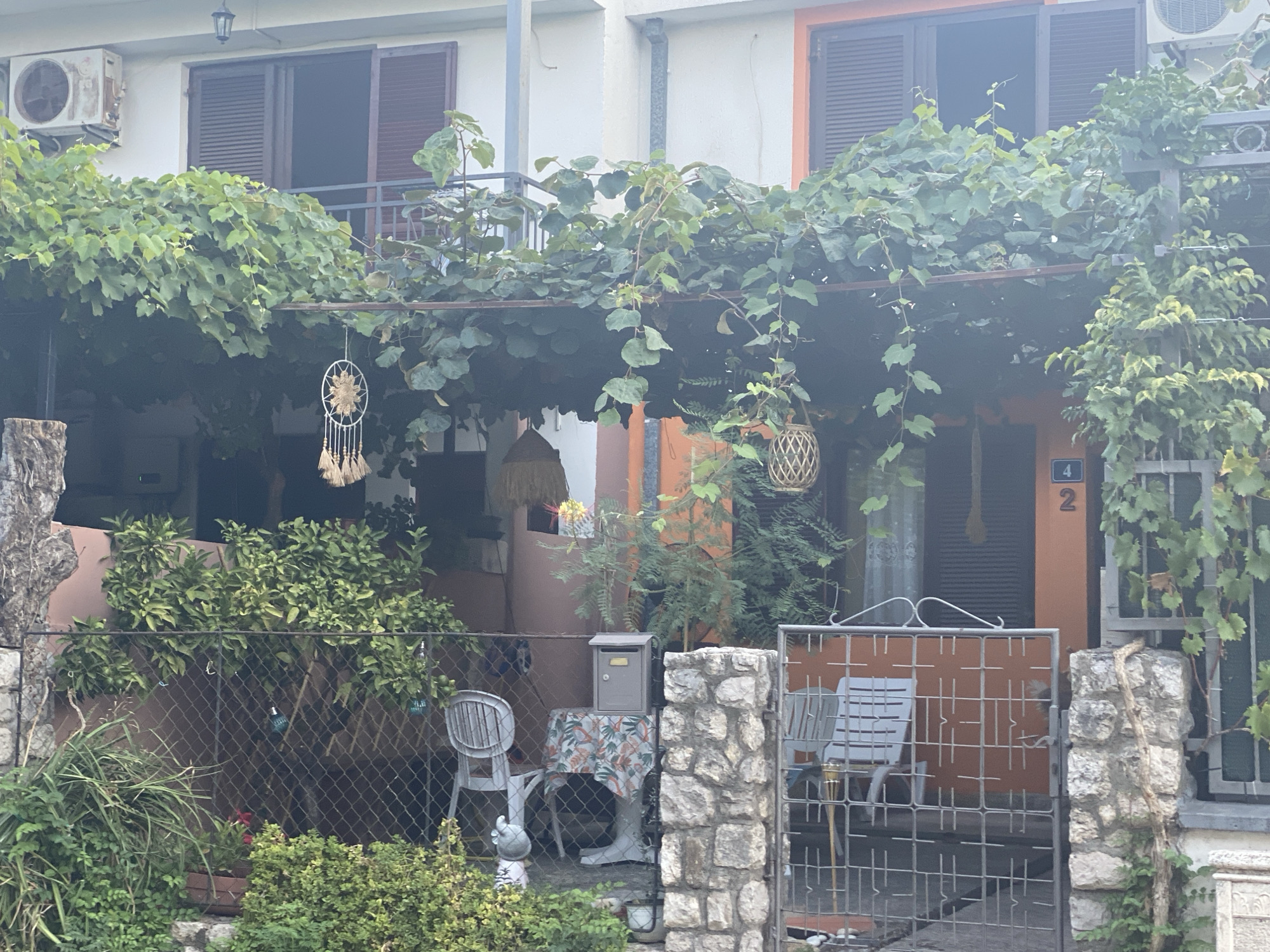 The image shows an outdoor scene in Sutomore, Montenegro. In the foreground, there is a fence and lush green vegetation. Behind the fence, there is a courtyard with several chairs and a table covered with a patterned tablecloth. A mailbox sits on the table. Beyond the courtyard, there is a building with orange and white facades, featuring windows with dark shutters and an air conditioning unit. Green vines hang over a covered patio area. A dreamcatcher is suspended from the patio. The overall impression is of a residential area with a focus on outdoor living space.