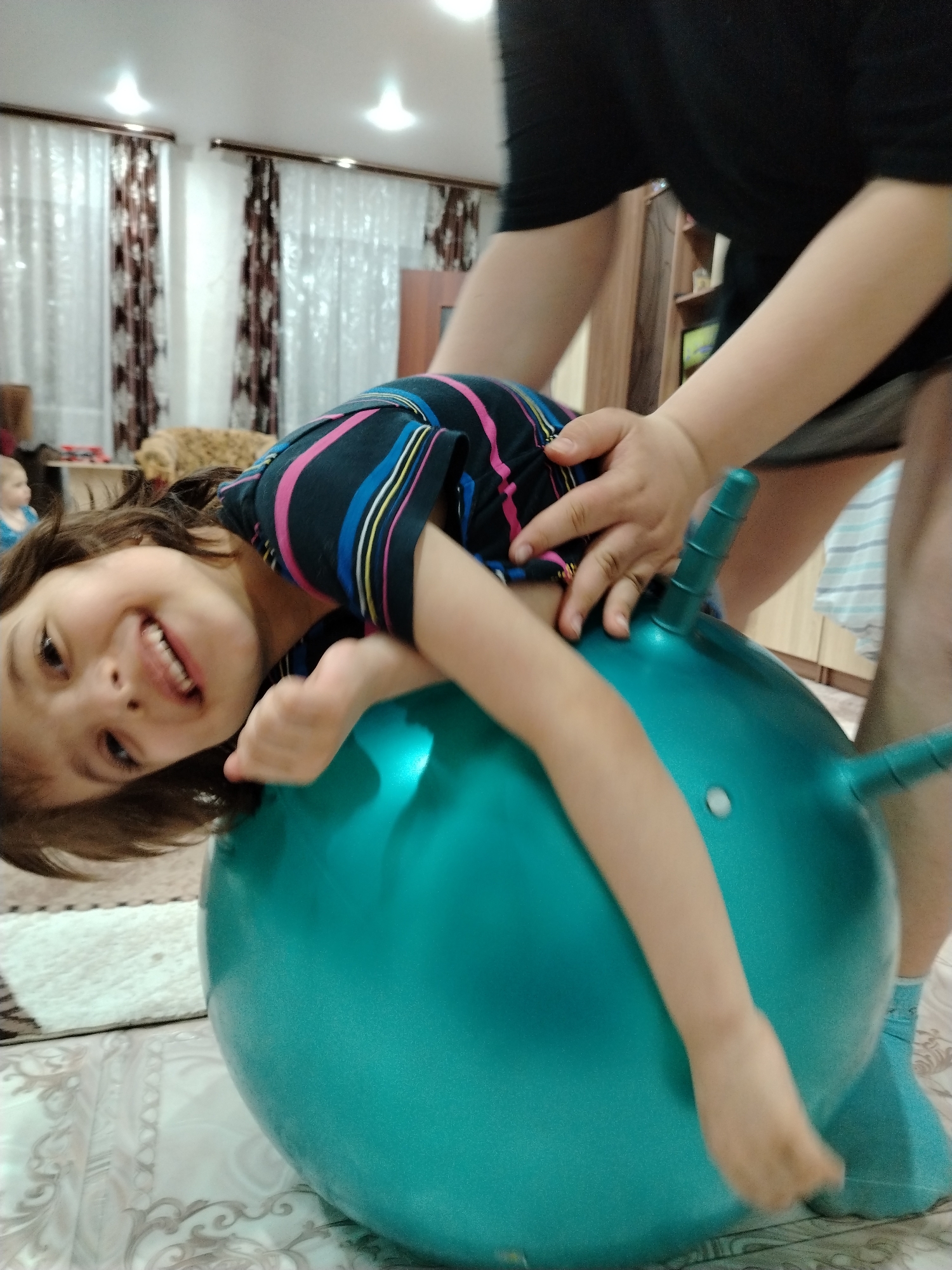 A child with brown hair and a striped shirt lies on top of an inflated, turquoise bouncy ball with handles. Their face is visible, showing a smile. An adult is positioned behind the child, holding the handles of the bouncy ball. The adult wears a black shirt and short skirt. The setting appears to be an indoor space with patterned curtains and a decorative floor. The location is believed to be in Большая Уса, Russia.