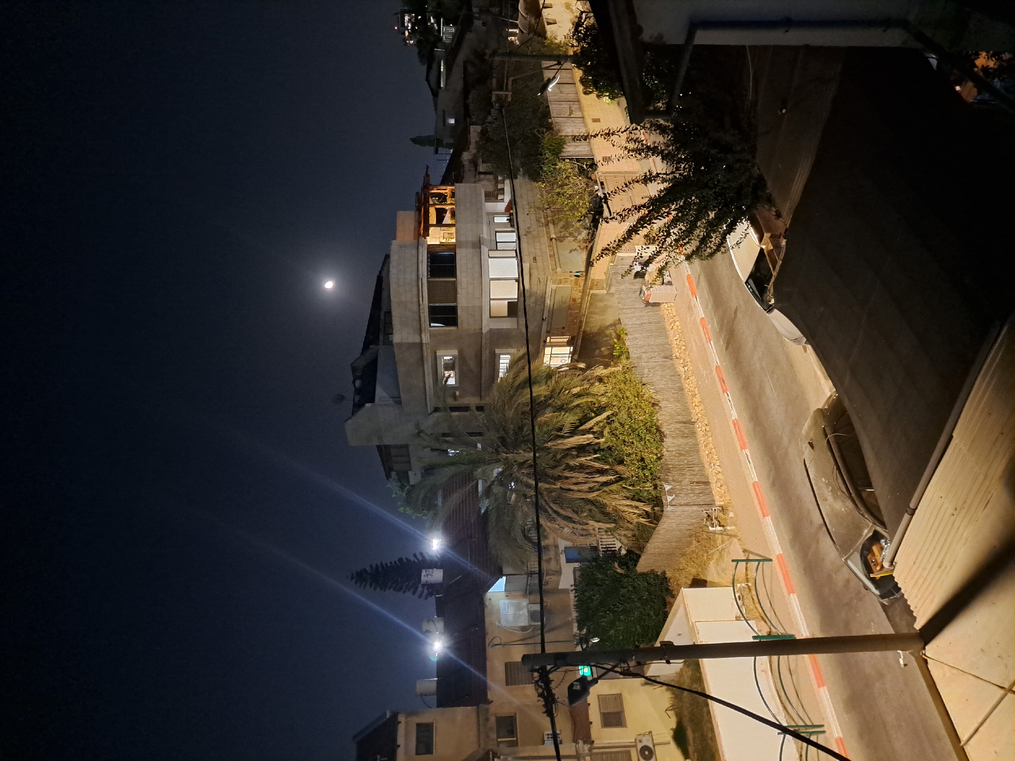 This is an outdoor, eye-level, long-exposure night photograph of a residential street in Ramat HaSharon, Israel. The scene is illuminated by artificial lights and moonlight. In the sky, a crescent moon is visible above the buildings. The street is paved and has a sidewalk with a red and white striped curb. On the street, there is a parked, dark-colored car with its doors open. A person wearing a yellow shirt is visible in the driver's seat of the car, holding a yellow object. The surrounding environment includes several multi-story buildings with windows, balconies, and rooftops. There is a large palm tree in the foreground, and various other trees and bushes. A wooden fence borders the property. Electrical wires and a utility pole are also present in the scene.