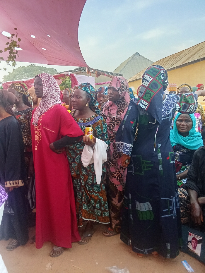 A group of women stand outdoors, dressed in colorful, patterned attire with head coverings. One woman holds a can and a white cloth. The setting appears to be a gathering or event, possibly a celebration, given the pink draped canopy above. Buildings are visible in the background. The ground is dirt. Some individuals are seated, and a bag with a portrait on it is on the ground.