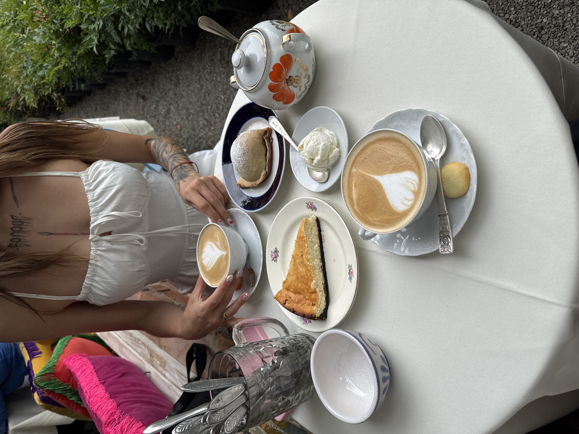 A woman with tattoos on her arm and torso, wearing a white dress, is seated at a table laden with desserts and coffee. The table is covered with a white tablecloth, and the scene appears to be outdoors, possibly at a cafe with a garden setting. In the background, rows of potted plants are visible, and the ground is covered in gravel, suggesting a location in Tbilisi, Georgia, known for its many charming outdoor cafes and gardens.
On the table, there are two cups of coffee with latte art, a slice of cheesecake, a pastry dusted with powdered sugar, and a scoop of ice cream. A decorative teapot, a small bowl, and cutlery are also present. The woman is holding one of the coffee cups, her hands adorned with rings and painted nails. The overall ambiance is relaxed and inviting.