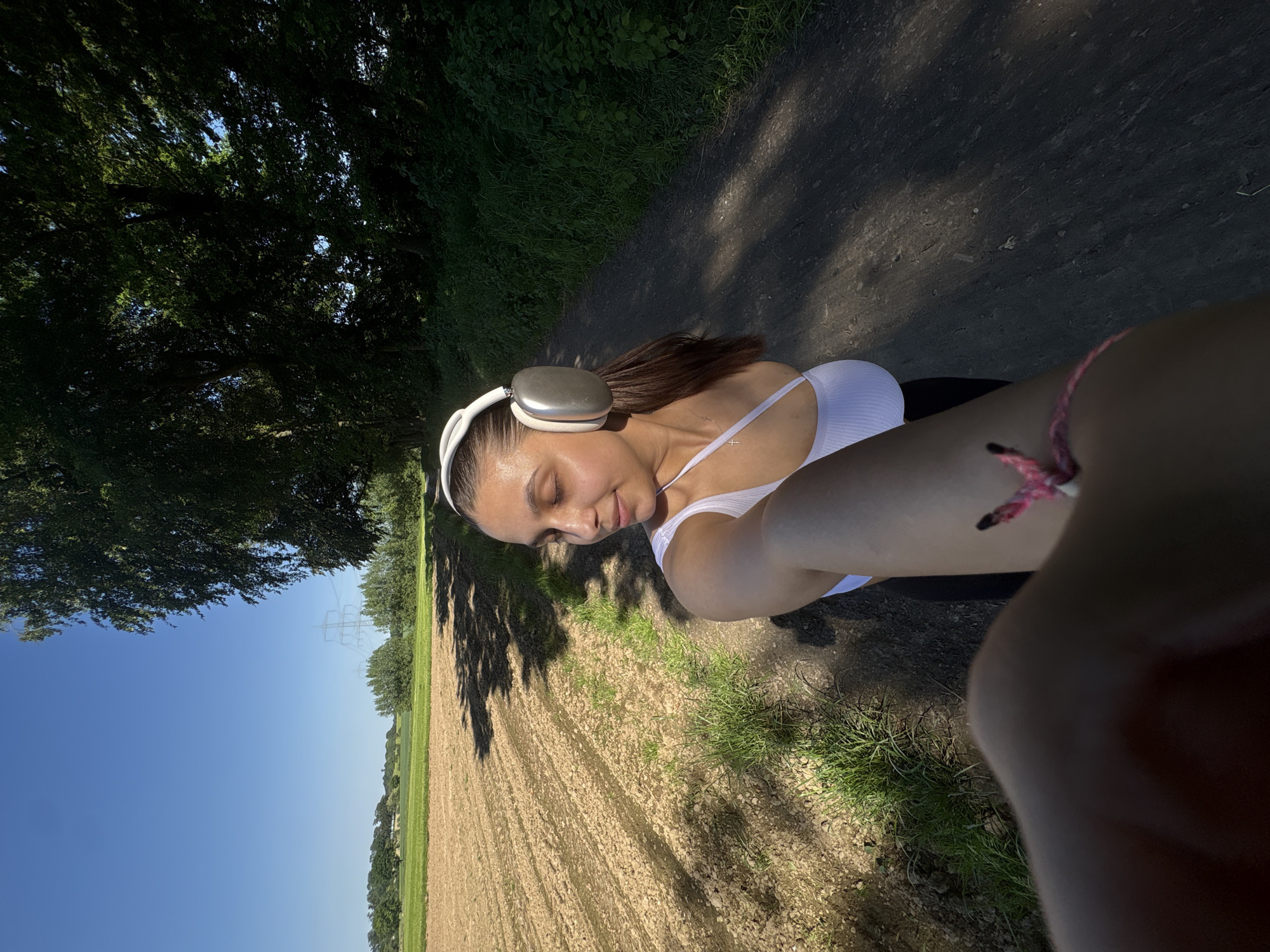 A selfie of a woman is taken in a rural area of Viersen, Germany. The subject is wearing white headphones, a white top, and dark bottoms. She has brown hair and is looking down. The background includes a dirt path, trees, a field, and a blue sky with visible power lines in the distance. The photo is taken during daylight with strong sunlight.