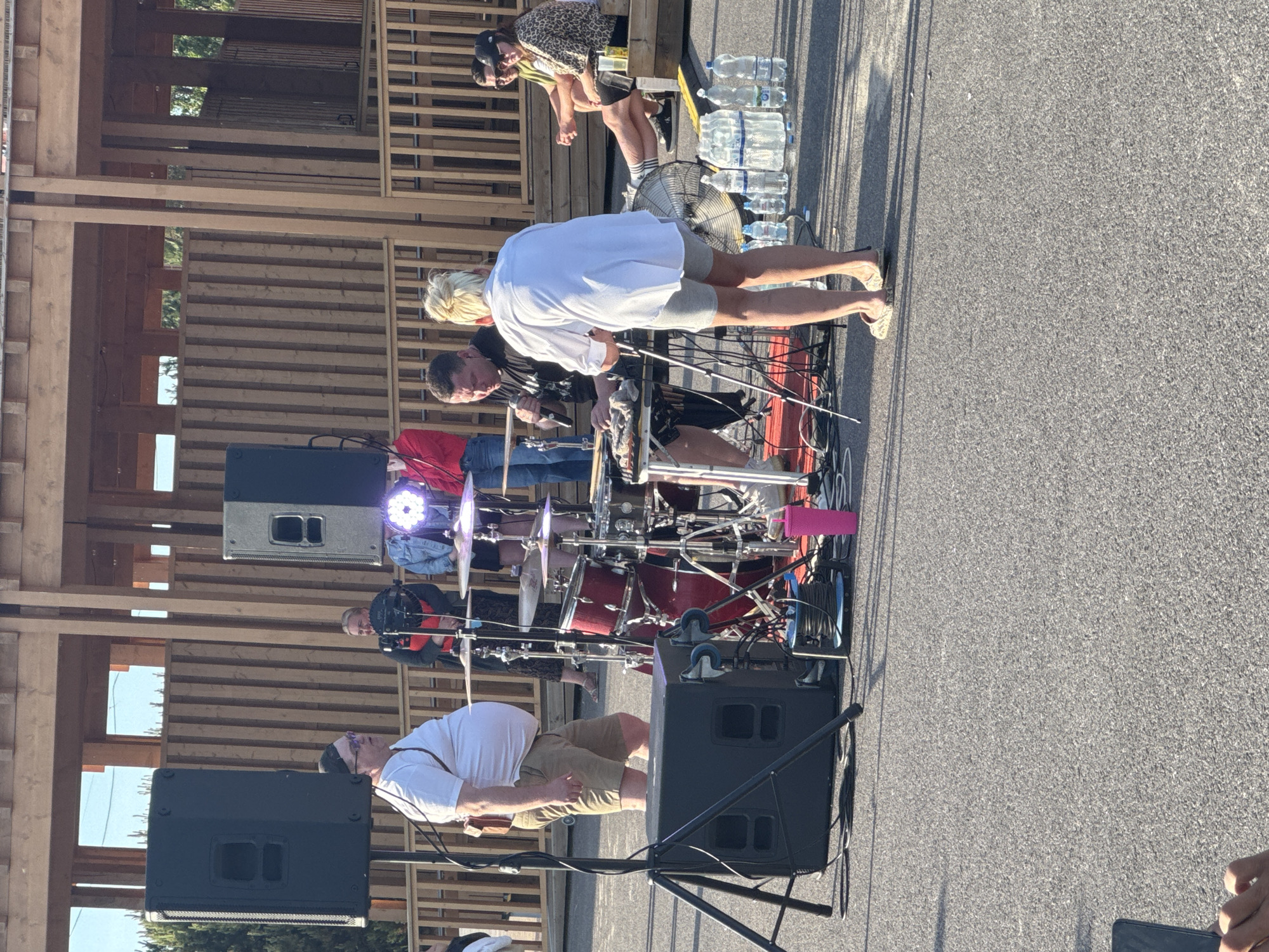 The image depicts an outdoor gathering in Tallinn, Estonia, likely at an event venue with a wooden structure in the background. In the foreground, a drum set is set up on a red mat. Two individuals are actively engaged with the equipment: a man wearing a black t-shirt is seated behind the drums, holding a microphone, and a person with blonde hair, wearing a white shirt and grey shorts, stands facing away from the camera, appearing to assist. To the left, a man in a white t-shirt and khaki shorts stands near a large speaker. Several people are visible in the background, some standing and observing. A row of water bottles is placed on the ground near a large fan. The ground surface is asphalt, and the lighting suggests it is daytime, possibly late afternoon due to the shadows.
