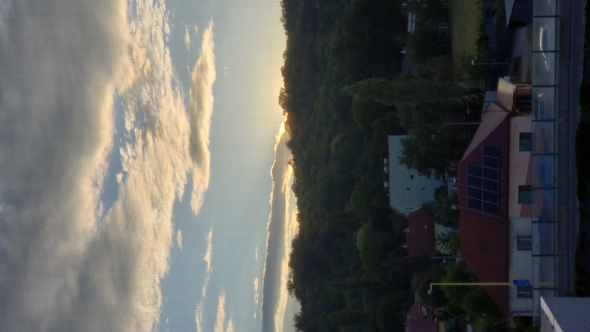 The image captures a sunset scene over a wooded hillside in Prešov, Slovakia. The sky is filled with a mix of clouds, with patches of blue visible and the sun's rays breaking through the clouds at the horizon, casting a warm glow. Below the sky, a dense green forest covers a hilly landscape. In the foreground, a residential area is visible. One house in the foreground has a dark red roof with an array of solar panels installed on it. Adjacent to this house, another building appears to be under construction or renovation, with exposed white wall sections. Further back, a light blue building stands among the trees. The scene is viewed through a window with a visible grid pattern.