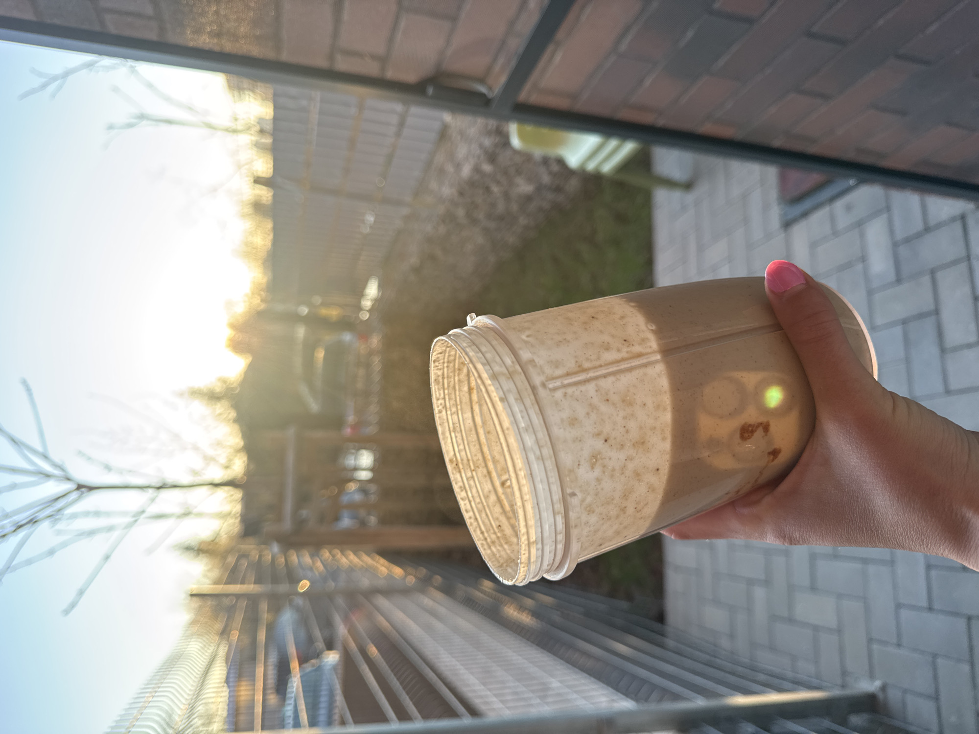 A person with pink nail polish is holding a blender cup with a light brown smoothie inside. The person is outdoors on a patio with gray pavers. There is a metal railing in the foreground. Behind the railing, there is a blurry view of trees and buildings. The light suggests it could be either dawn or dusk. The image does not contain enough information to determine if it is in Sofia, Bulgaria.