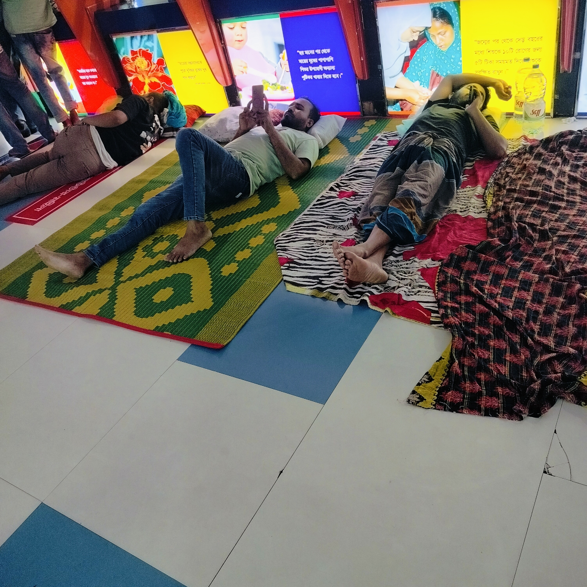 In a public space, potentially a transit area in Dhaka, Bangladesh, three individuals are seen lying down on makeshift beds. The man in the middle is using a smartphone, while the other two individuals appear to be resting or sleeping. The area features illuminated advertising boards above. There is a focus on providing healthy baby food.
