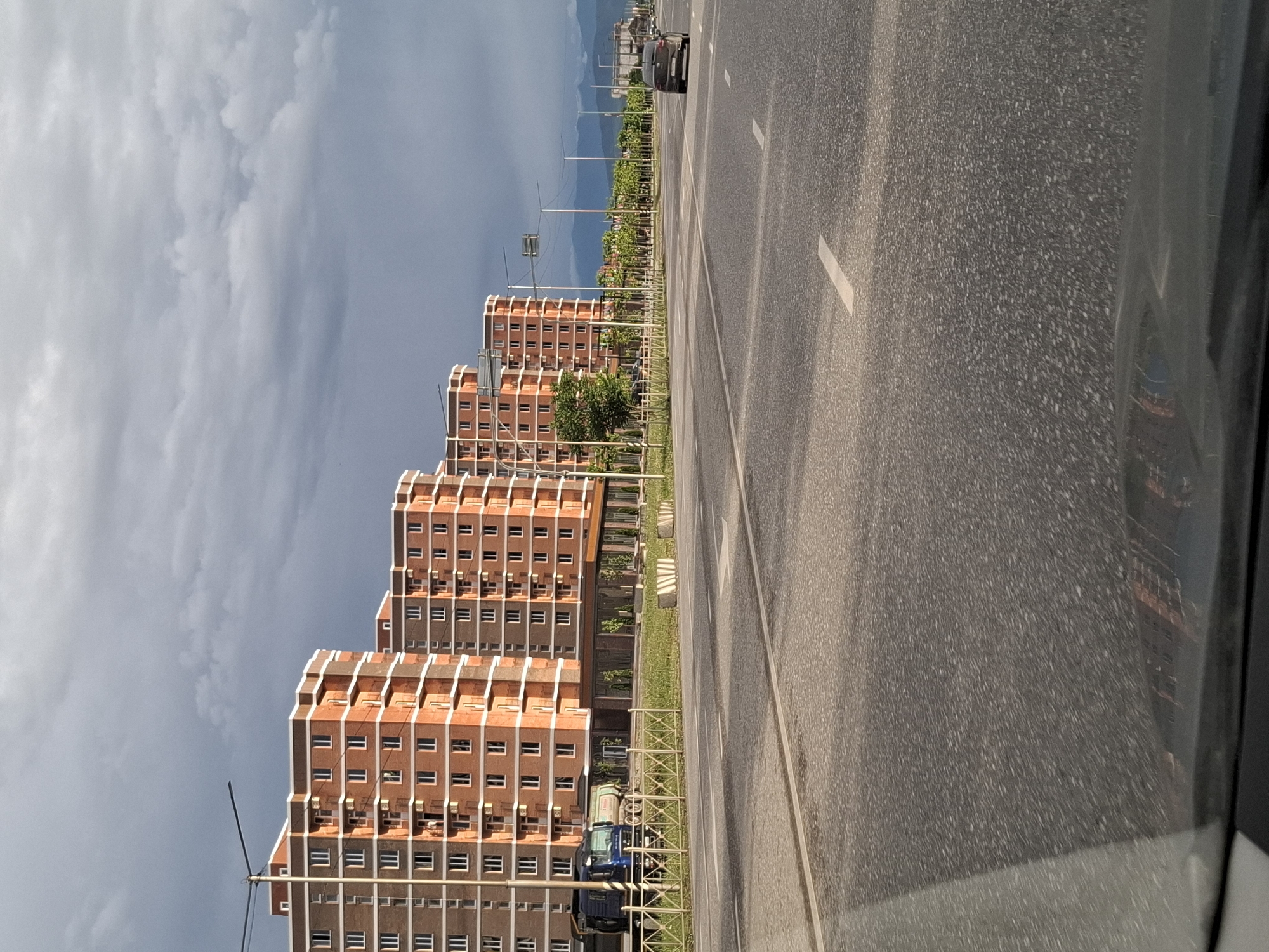 The image shows a street scene with modern, multi-story buildings made of red brick and gray concrete. The buildings are tall and have many windows. In the foreground, there is a paved road with white lane markings. A dark-colored van is parked on the side of the road. The scene appears to be in a developed urban or suburban area. The sky is overcast with gray clouds, suggesting a possibly late afternoon or early evening time. The presence of trees and a sidewalk area between the buildings and the road indicates a planned urban environment. The image is taken from a moving vehicle, as suggested by the slight blur and the perspective. The location is identified as Urus-Martan, Russia.