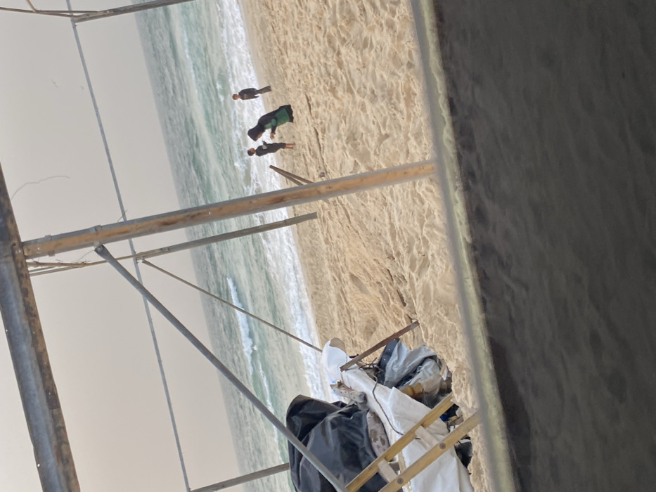 A beach scene is viewed through a construction framework. The beach has beige sand and gentle waves. Three figures are standing on the shoreline. Closer to the viewer, the construction site holds equipment and materials, including a ladder, white tarps, and various containers, suggesting ongoing work or temporary setup. The photograph's perspective is low, looking out towards the beach.