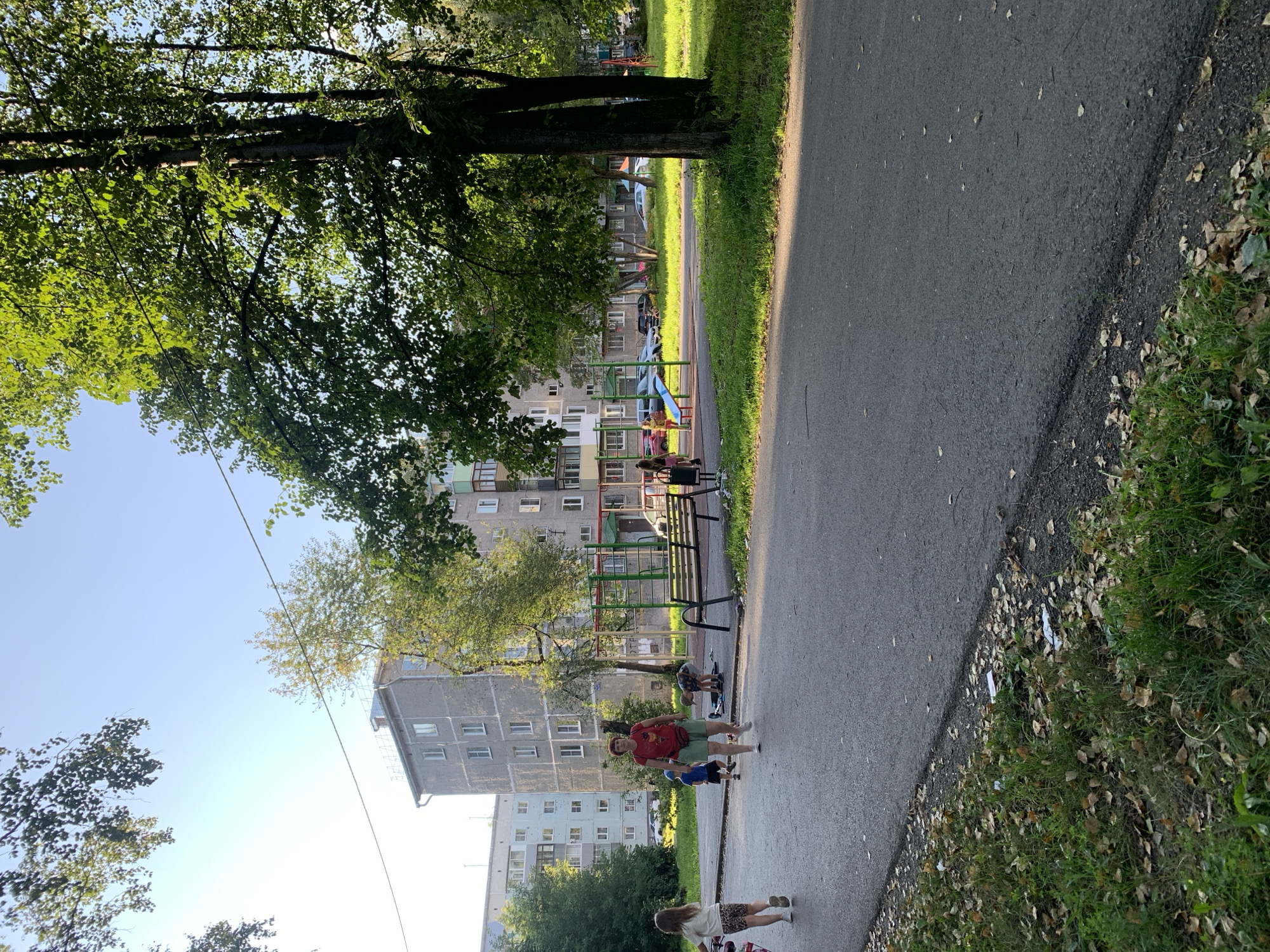 The image depicts a residential courtyard in Prokopyevsk, Russia, on a clear day. A paved walkway runs through the foreground, with a strip of grass and fallen leaves bordering it. Beyond the walkway, a playground area is visible, featuring a wooden bench, swings, and climbing structures. Several people are present in the scene. A woman in a red t-shirt and green shorts walks across the walkway, while a young girl in a white shirt and patterned shorts is to her left. Two young boys and a child on a scooter are near the playground. In the background, multi-story apartment buildings, typical of Soviet-era architecture, line the courtyard, interspersed with trees. Cars are parked along the street in front of the buildings. The overall scene captures a moment of everyday life in a Russian urban environment.