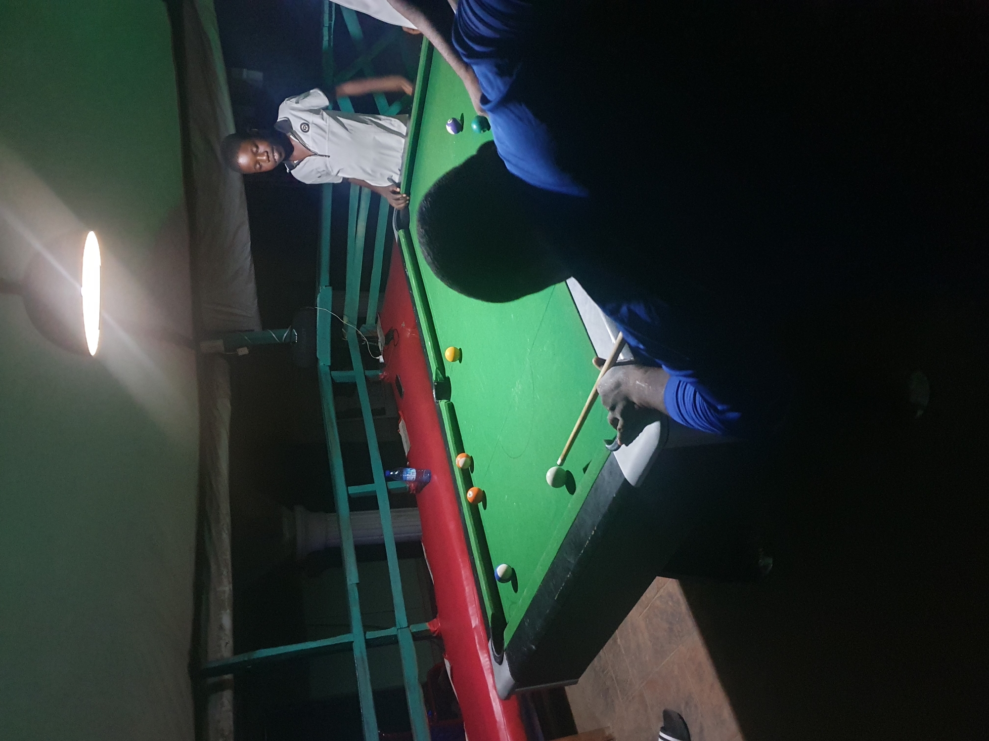 The image depicts two individuals in a dimly lit room, engaged in a game of billiards. A young person in a white uniform, possibly a school uniform, stands by the green billiard table, looking towards the camera with a neutral expression. Another individual, mostly in silhouette due to the lighting, is actively playing the game, holding a cue stick and aiming at a white ball. Several billiard balls, including solid and striped ones, are scattered across the table.
The setting appears to be a recreational area, possibly an indoor games room or a lounge, with the presence of a billiard table and a bright overhead light fixture illuminating part of the scene. The background is dark, suggesting it might be nighttime or a poorly lit environment. A red railing runs along the side of the billiard table, and a green metal structure, possibly a railing or part of the furniture, is visible in the background. A plastic bottle of water is placed on the railing.
The overall mood is relaxed and focused on the game. There are no overt emotional displays. The location context provided is Lokoja, Nigeria, although there are no specific visual cues in the image that definitively confirm this location. The time of day is implied to be evening or night due to the low lighting.